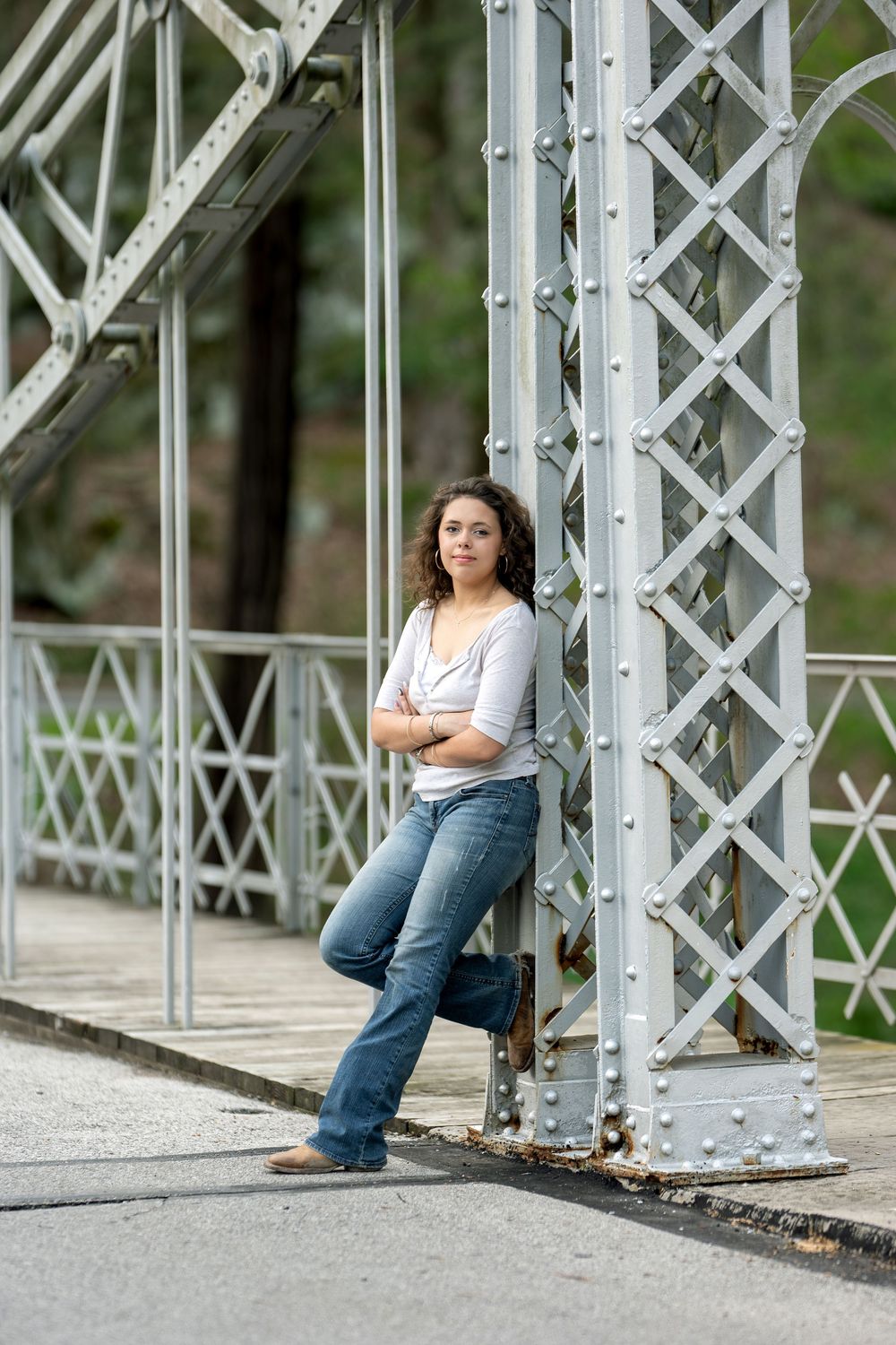 Young woman in jeans leaning against a metal bridge pillar with arms crossed in an outdoor setting.