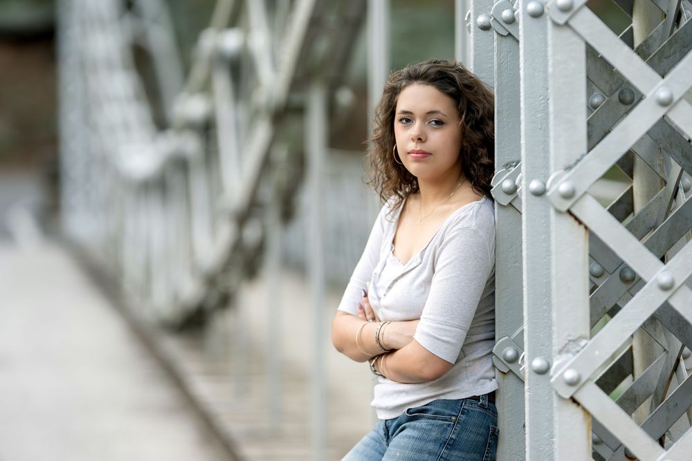 Young woman with curly hair standing with arms crossed against a metal bridge with a serious expression.