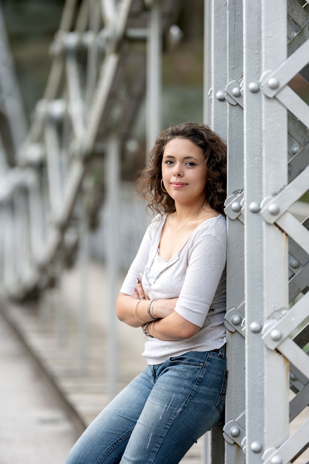 Curly-haired woman leaning against metal bridge support with arms crossed.