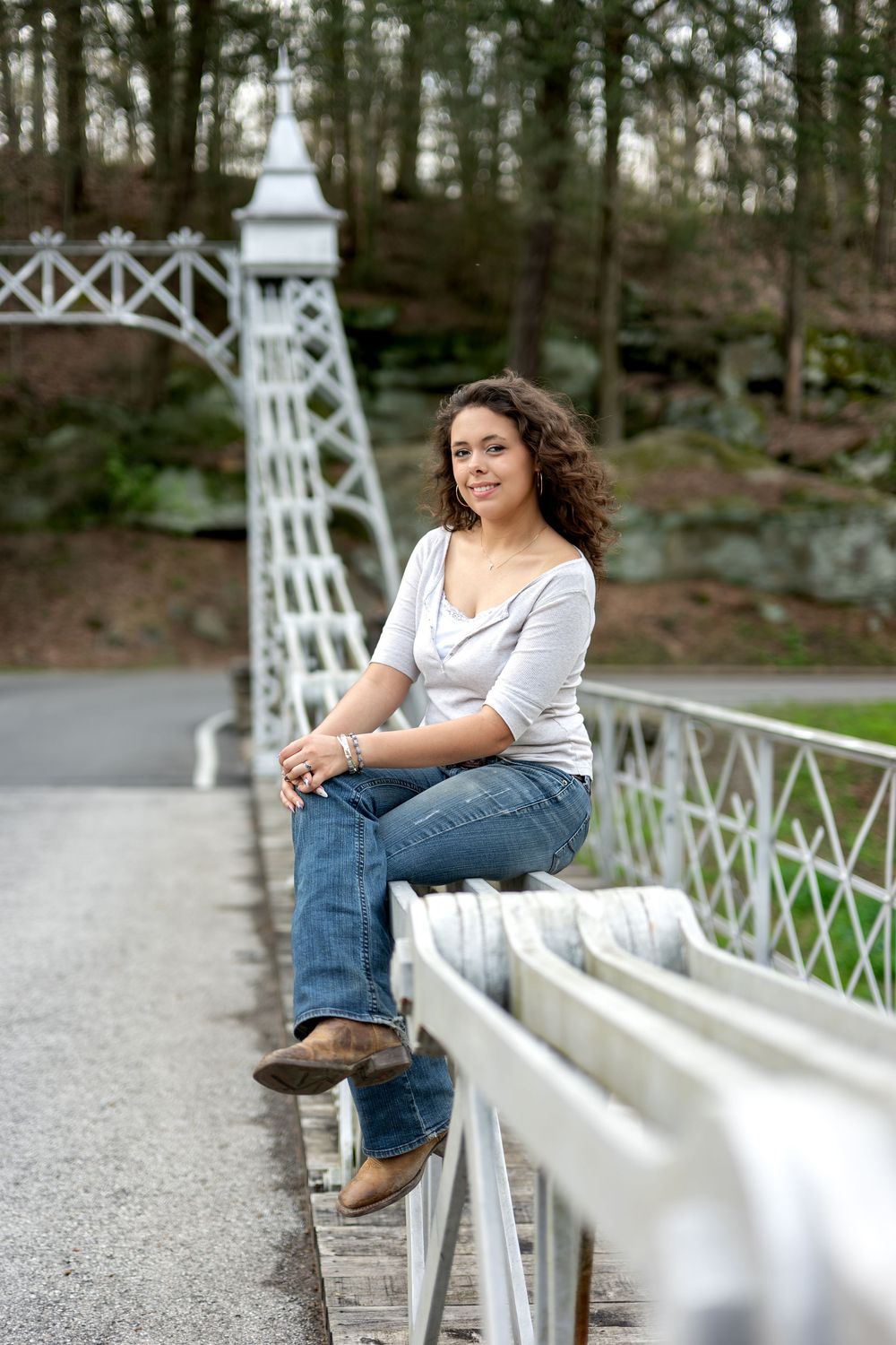 Young woman sitting on white metal bridge railing in wooded park.