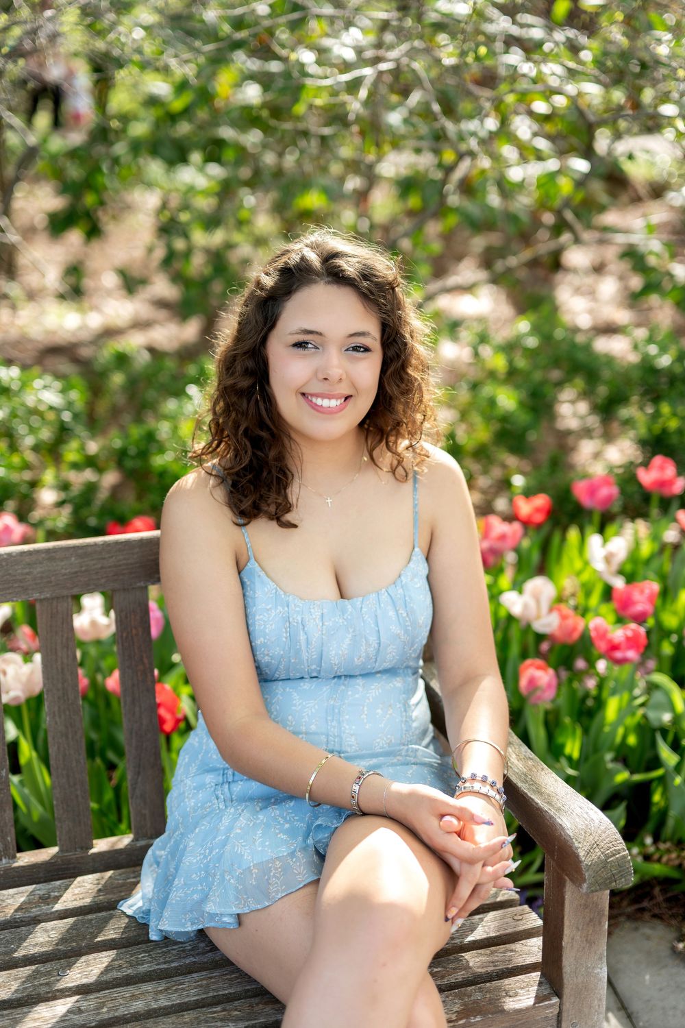 Young woman in a blue sundress sitting on a wooden bench surrounded by colorful tulips in bloom.