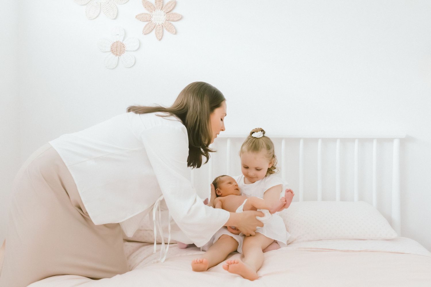 Mother and young daughter bonding with newborn baby on white bed in bright nursery room.