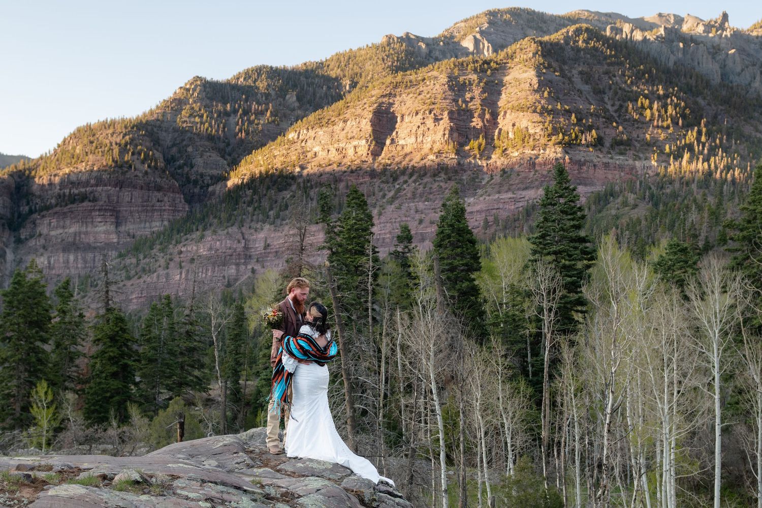 Couple sharing romantic embrace on rocky mountain overlook with evergreen trees and red cliffs at golden hour.