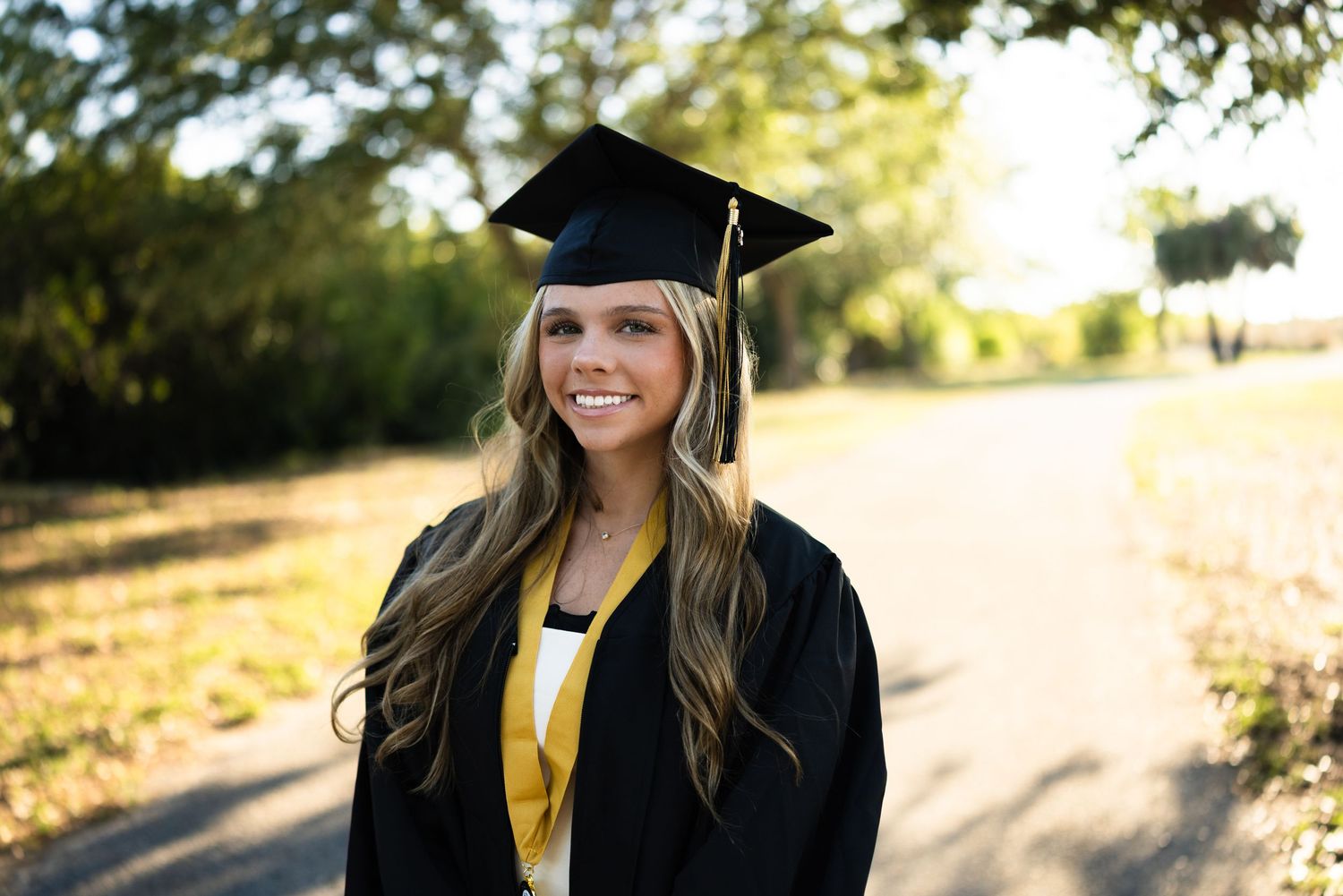 Smiling young woman in black graduation cap and gown with gold accents poses outdoors on a sunny day.