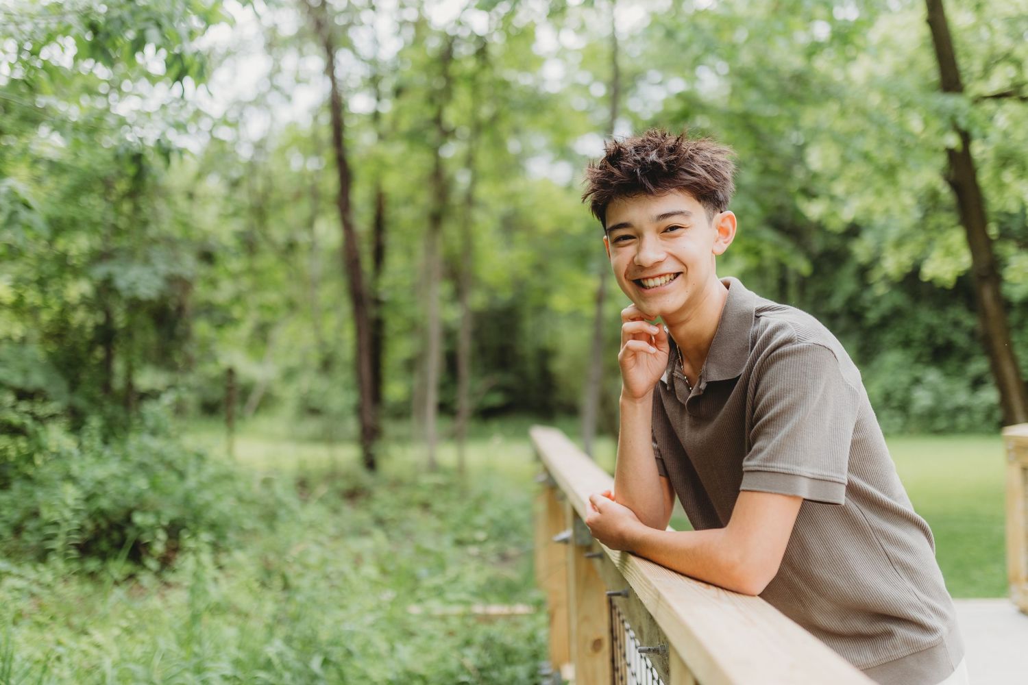 Smiling teenage boy leaning on wooden bridge railing in lush green park setting.