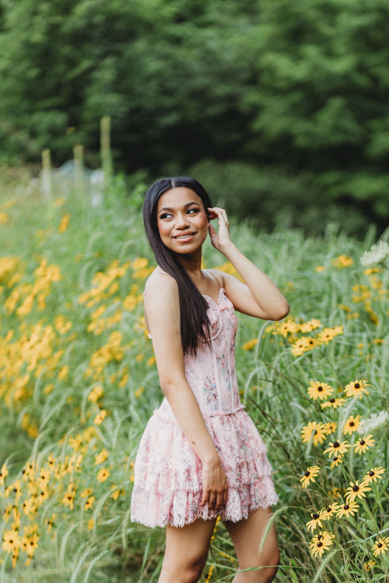Young woman in pink floral dress posing among yellow wildflowers and lush green foliage outdoors.
