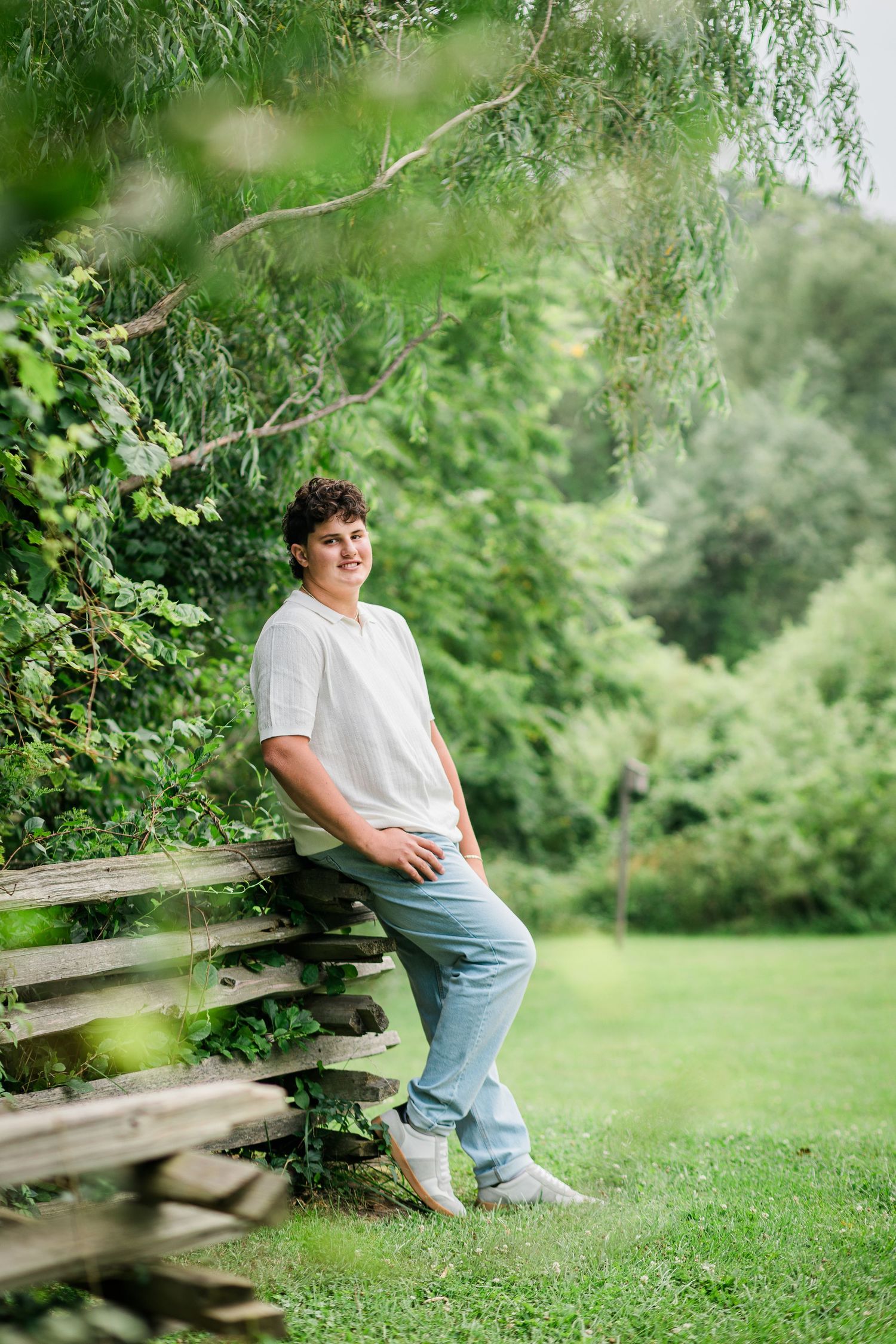 Teen boy in white t-shirt and jeans leaning on rustic wooden fence surrounded by lush green trees.