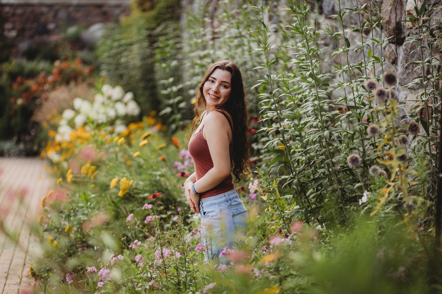 Young woman in red top and jeans smiling in a lush garden surrounded by colorful wildflowers and greenery.