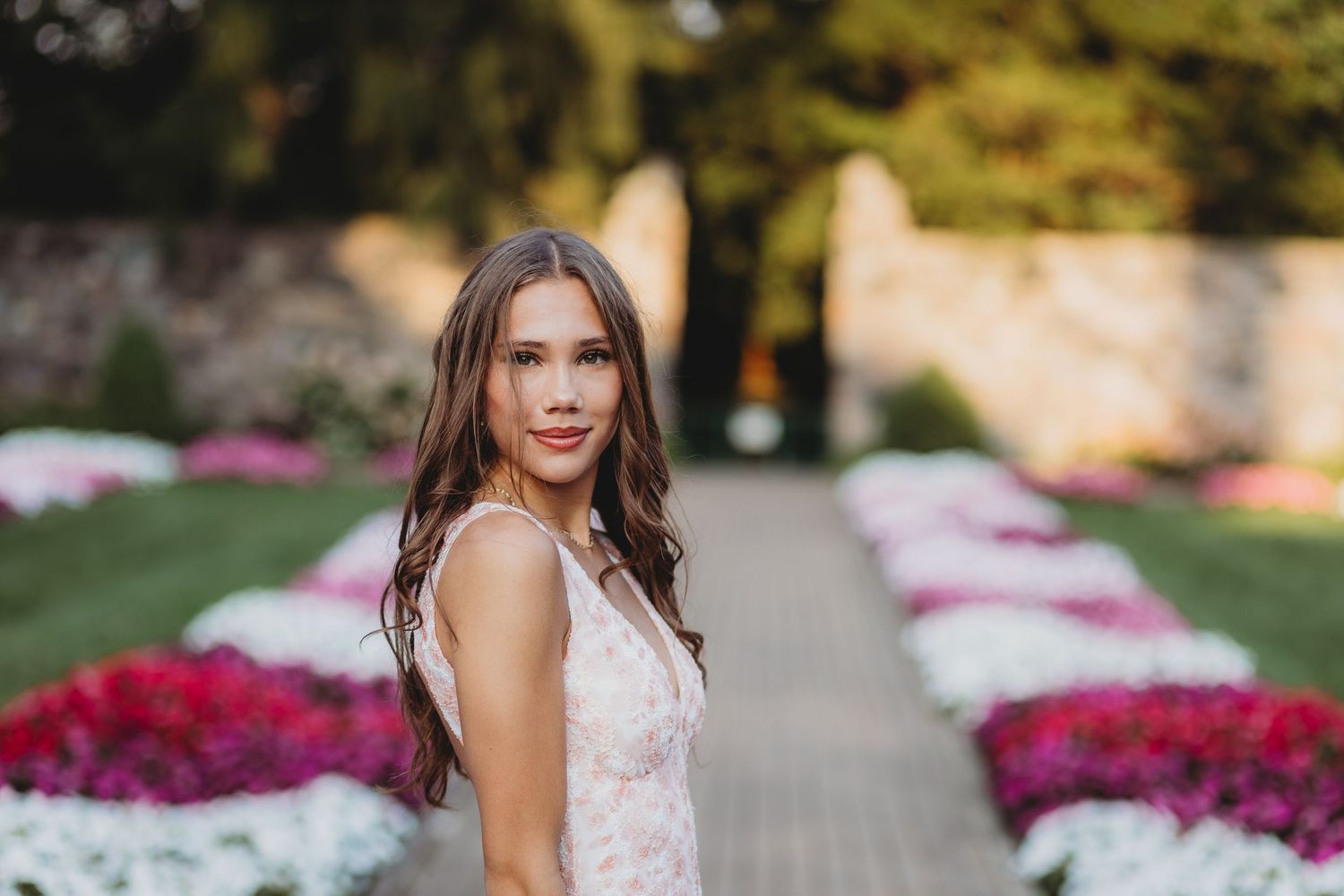 Young woman in pink lace dress poses in garden with colorful flower beds and stone pathway.