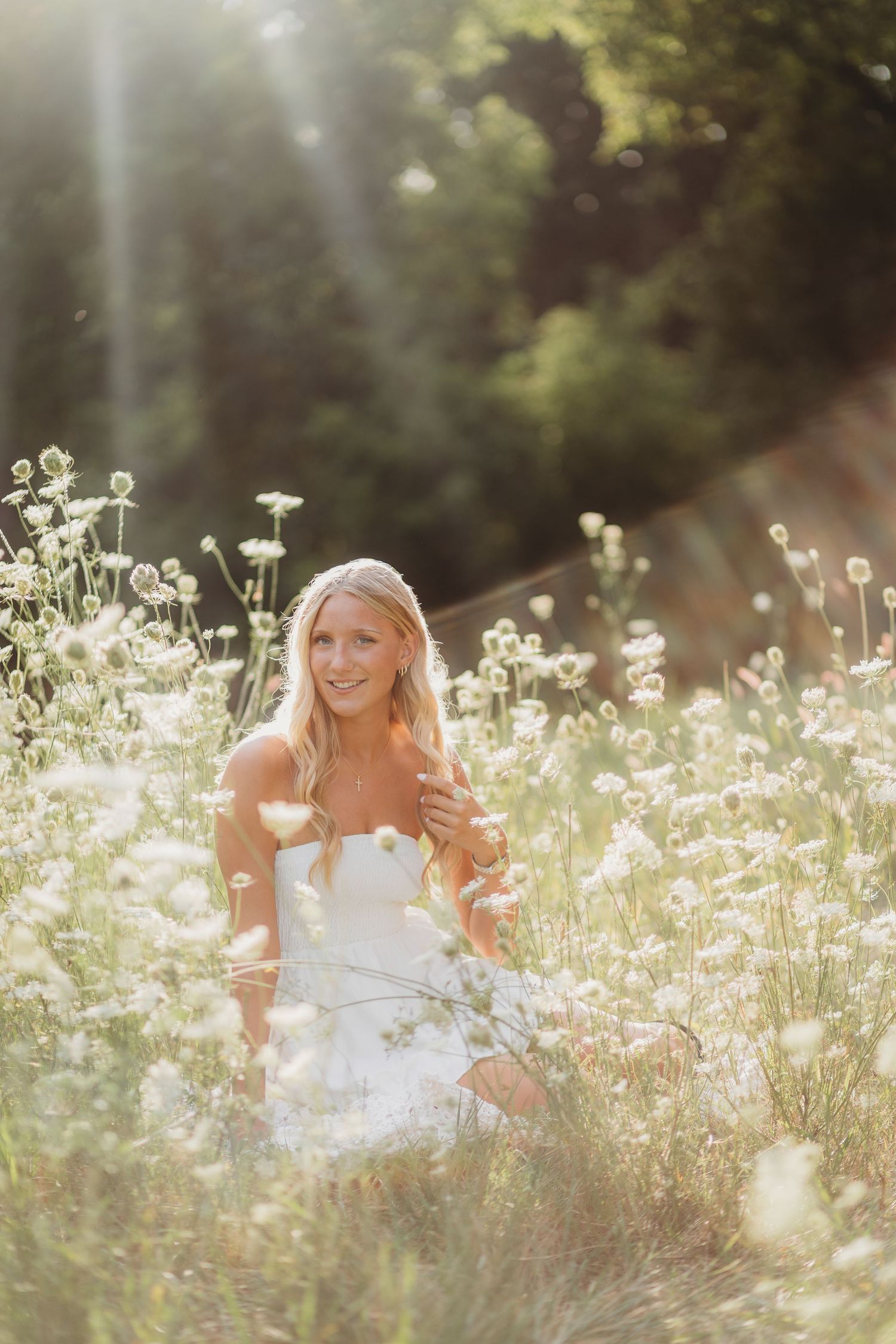 Smiling blonde woman in white strapless dress sitting in sunlit wildflower meadow with soft bokeh background.