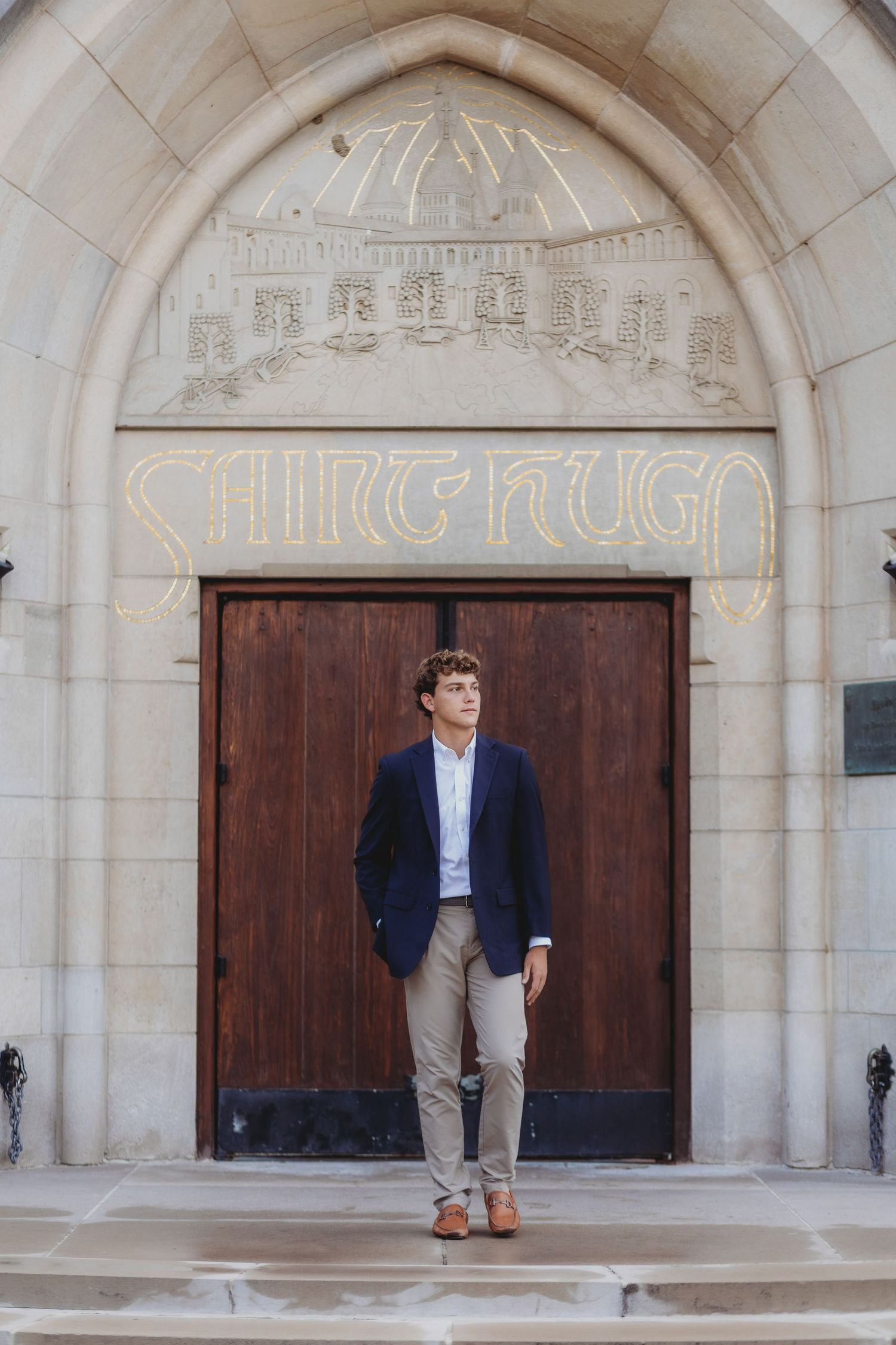 Young man in navy blazer and khakis stands before ornate stone archway of Saint Hugo church wooden doors.
