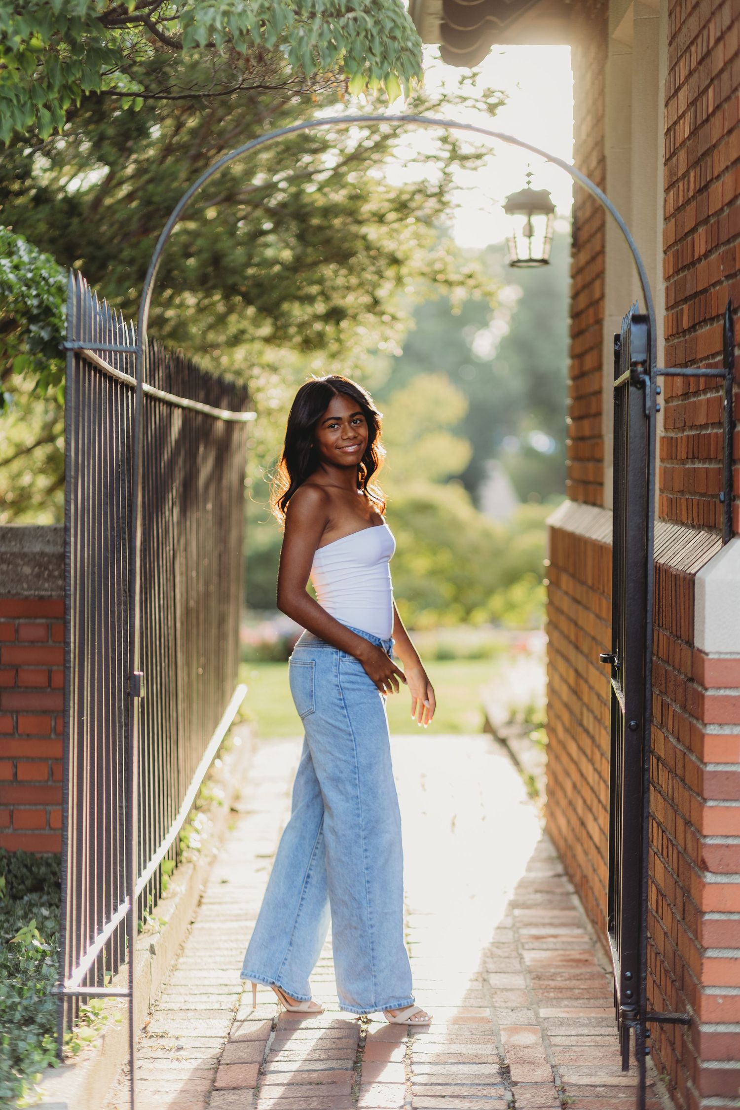 Young woman in white tube top and wide-leg jeans posing by an arched iron gate on a sunny day.