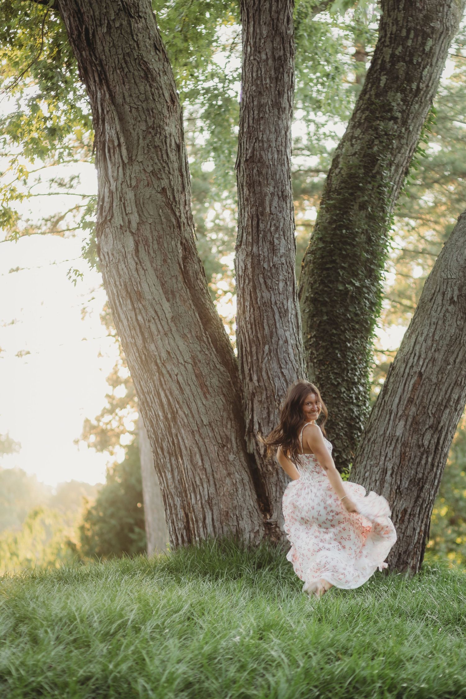 Woman in floral dress twirling near large trees in golden sunlight on lush green grass.