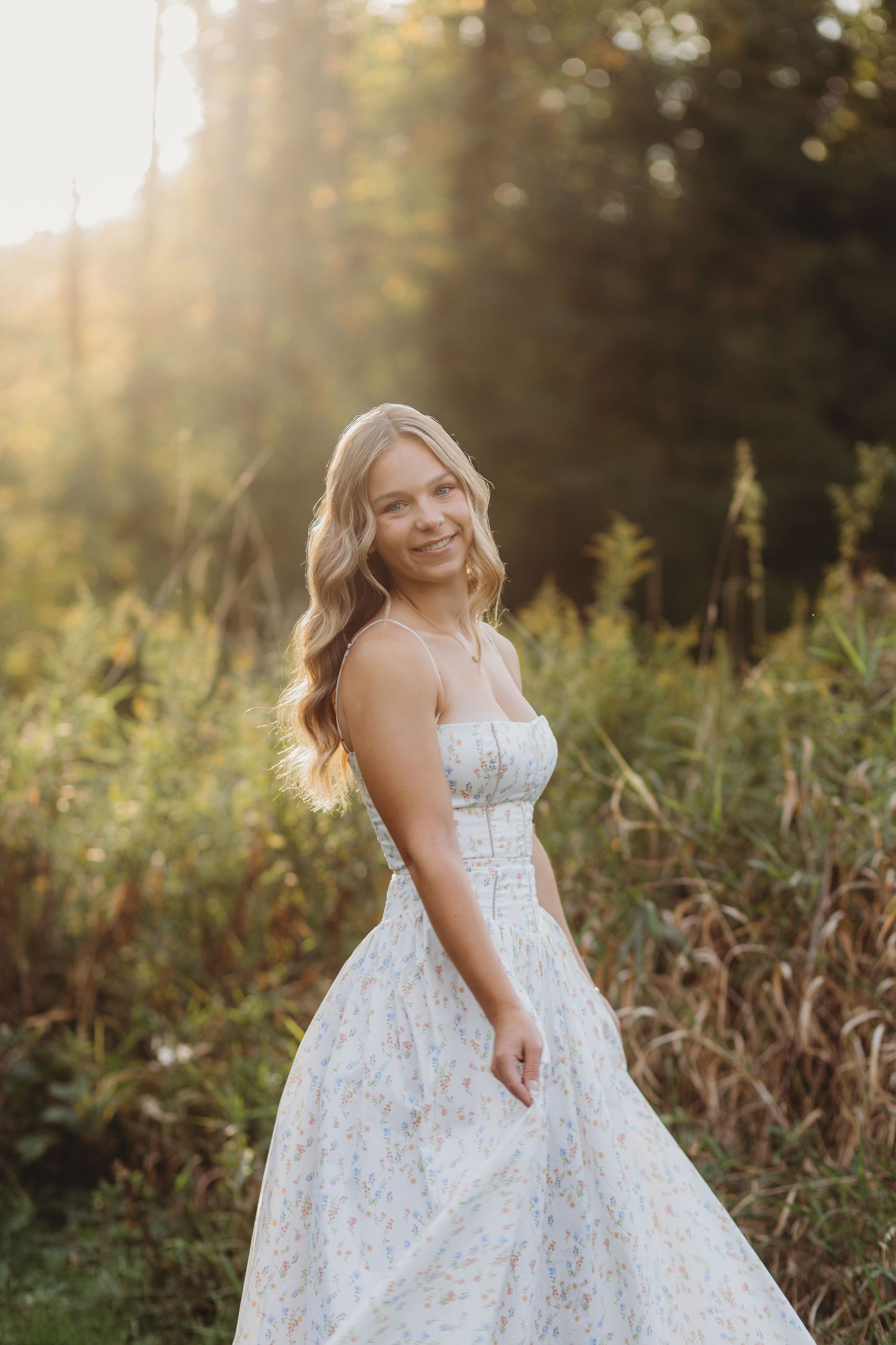 Young blonde woman in a floral sundress smiling outdoors in golden hour sunlight with lush greenery background.