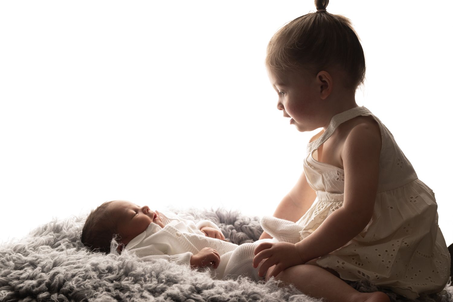 A toddler girl in a lace romper gazes lovingly at a sleeping newborn baby lying on a fluffy gray blanket.