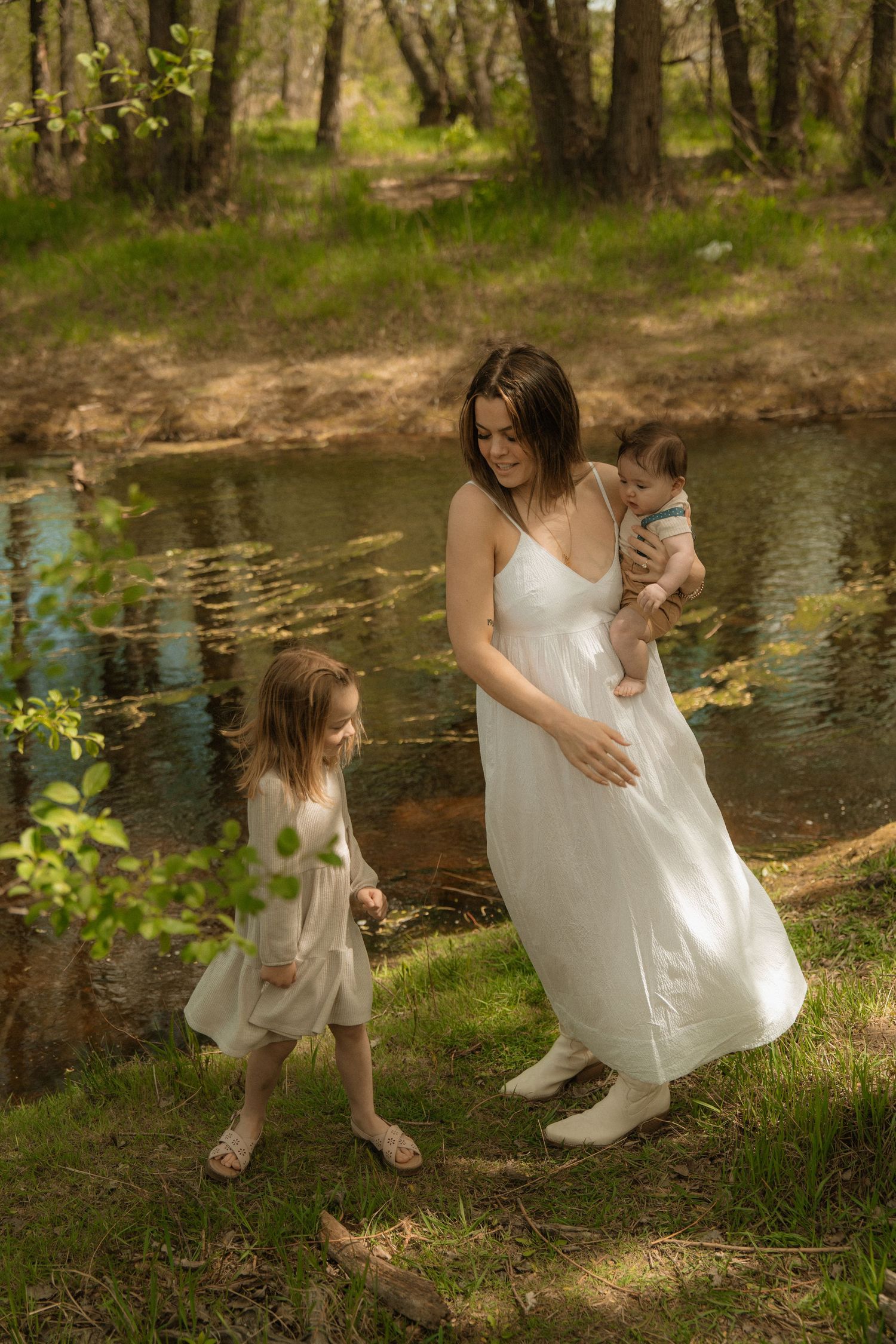 Family of three walking in golden field near rustic barn, man spinning woman in white dress while child holds flowers.