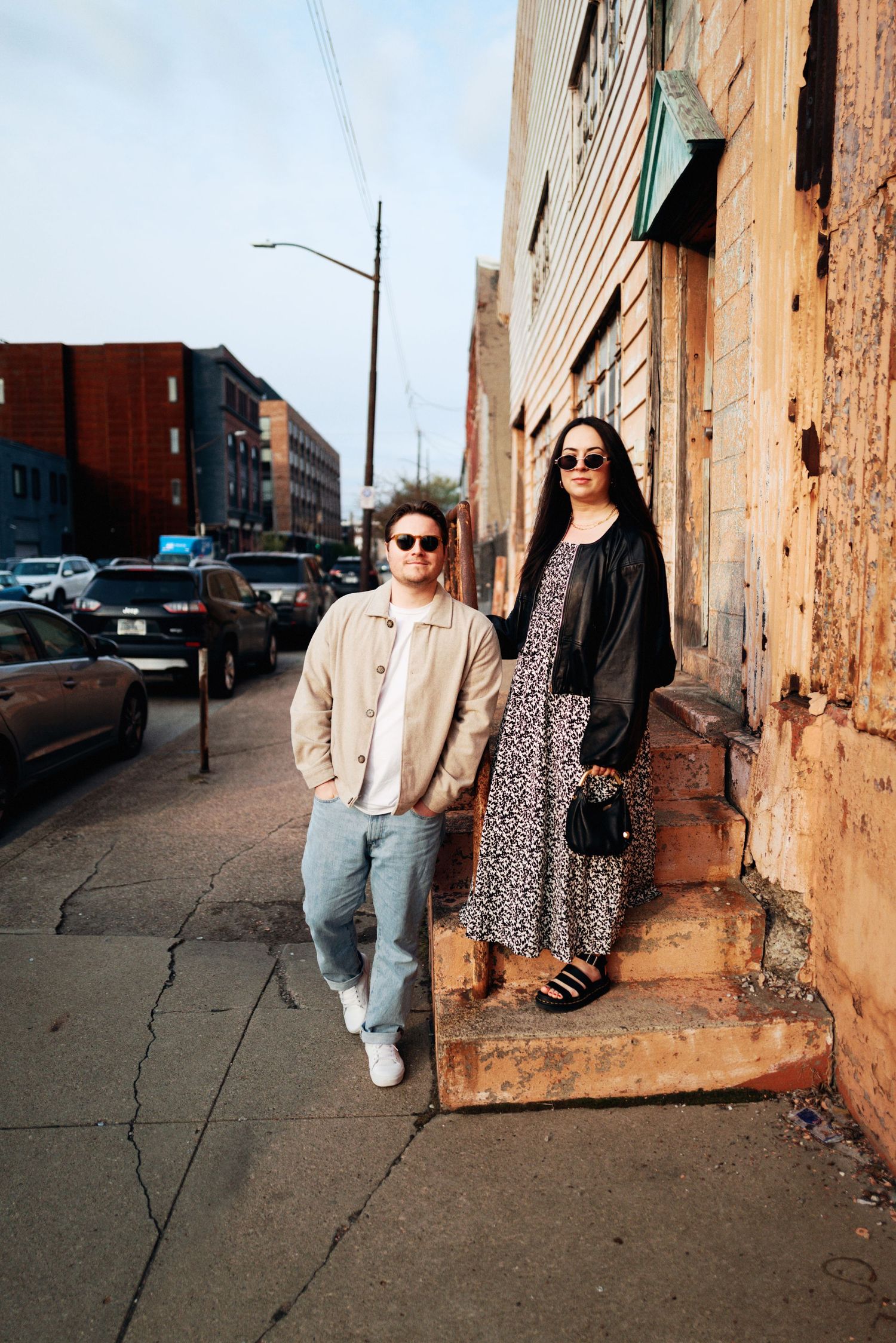Two stylish friends in sunglasses pose on urban sidewalk steps beside a weathered brick building on a sunny day.