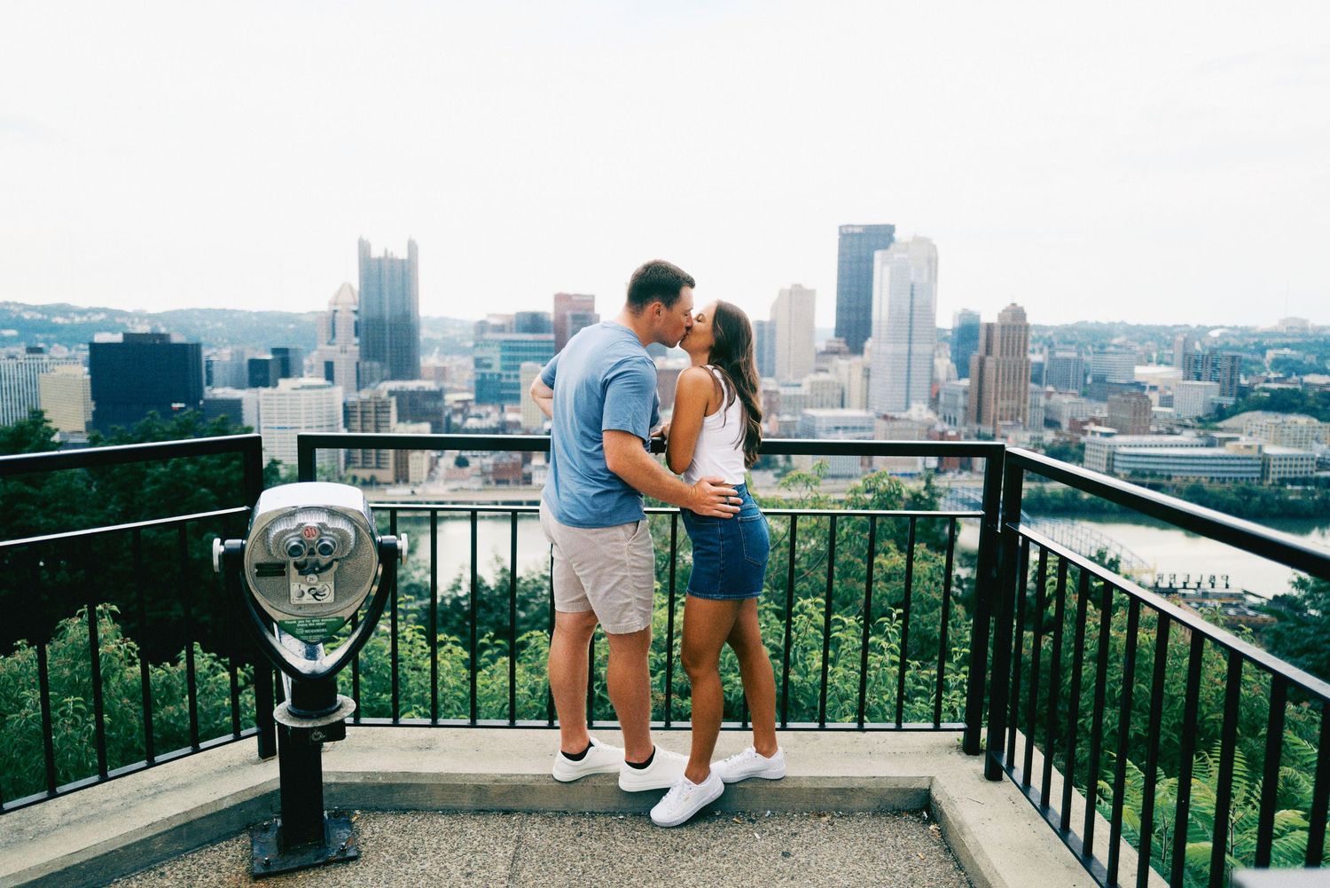 Couple kissing on an overlook with Pittsburgh city skyline and lush greenery in the background on a sunny day.