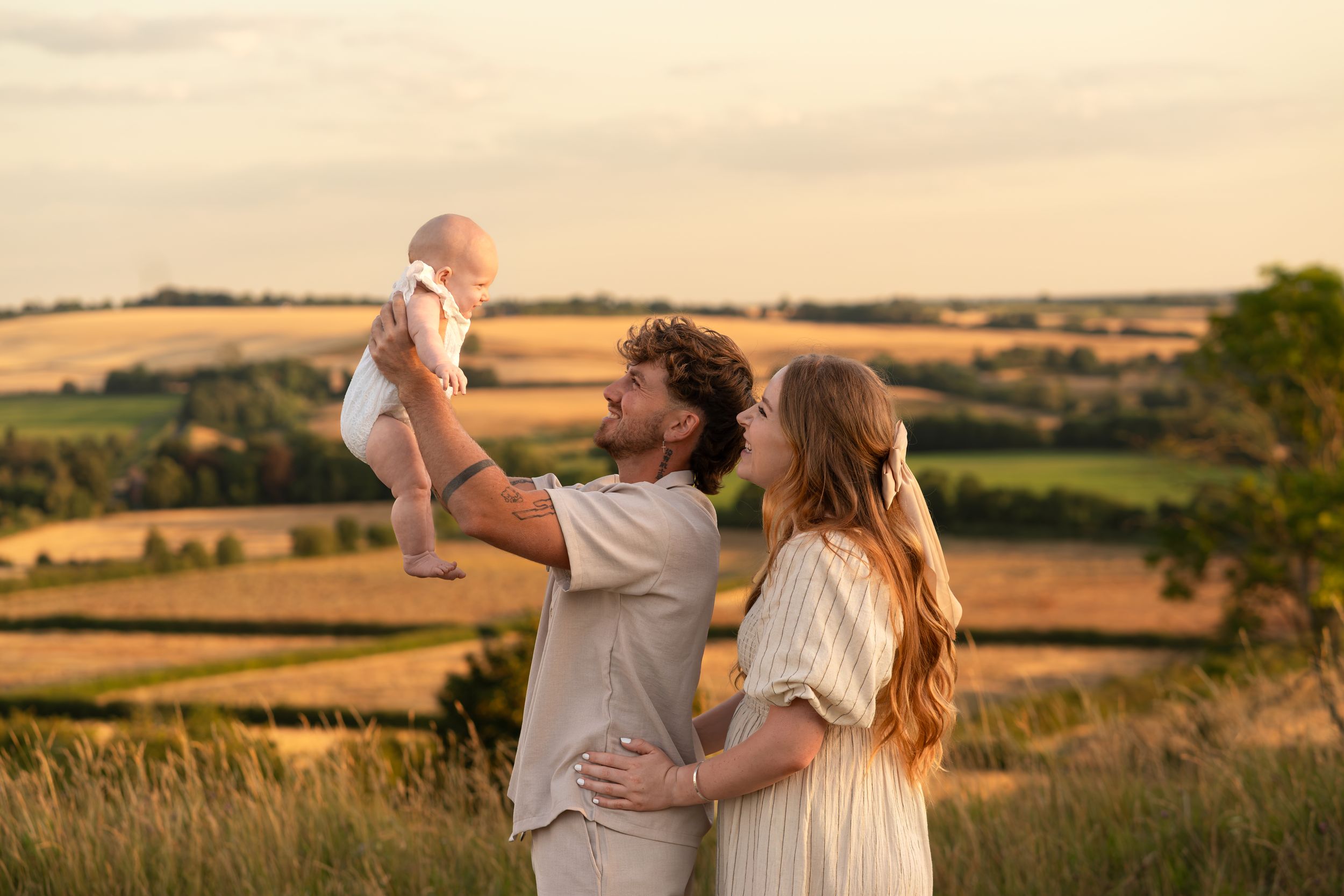A couple holds their baby outdoors at sunset with golden fields in the background.