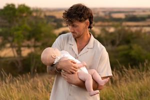A young man with curly hair tenderly holds a newborn baby outdoors in a golden field at sunset.