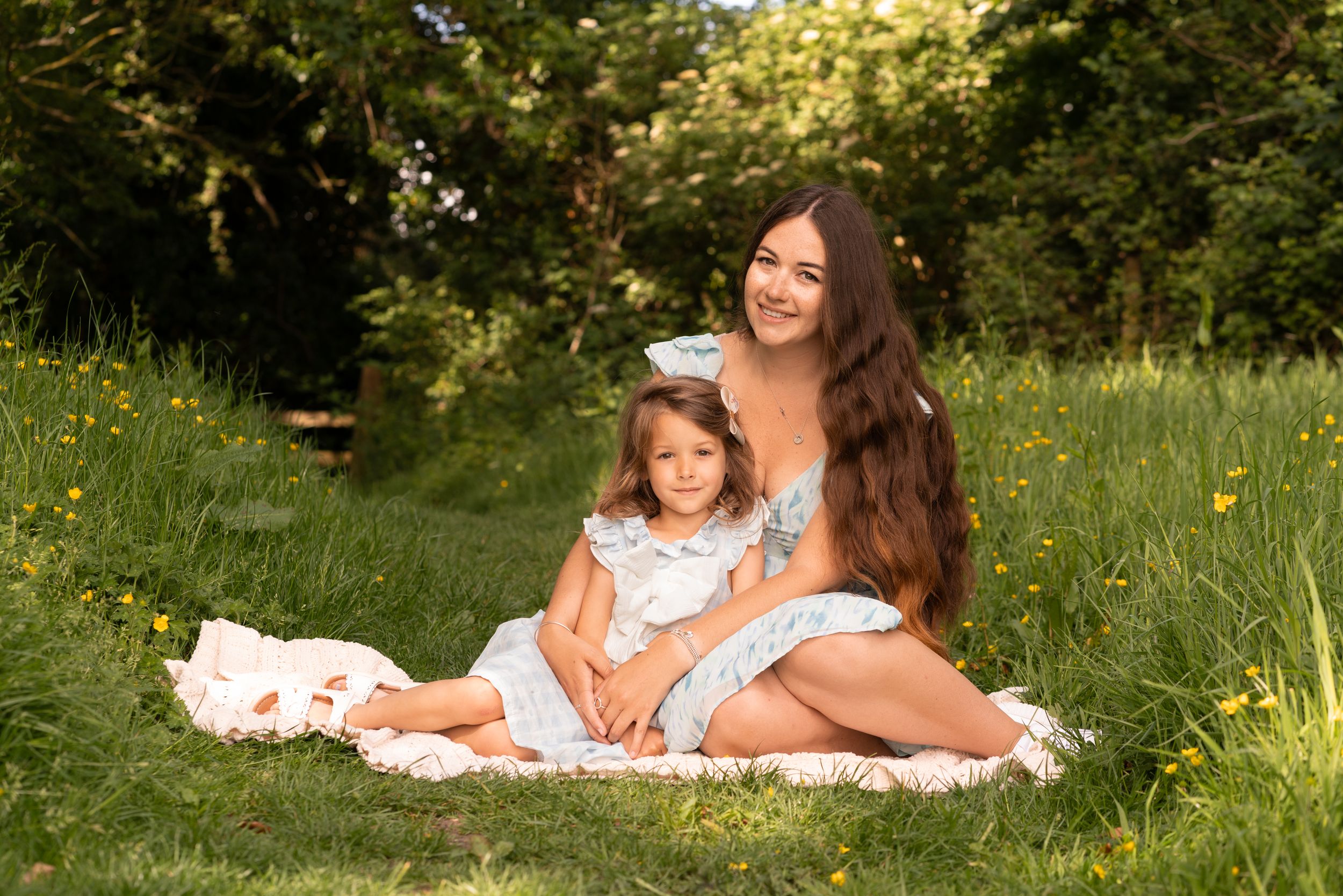 A mother and young daughter in matching white dresses sit together on a blanket in a sunny green meadow.