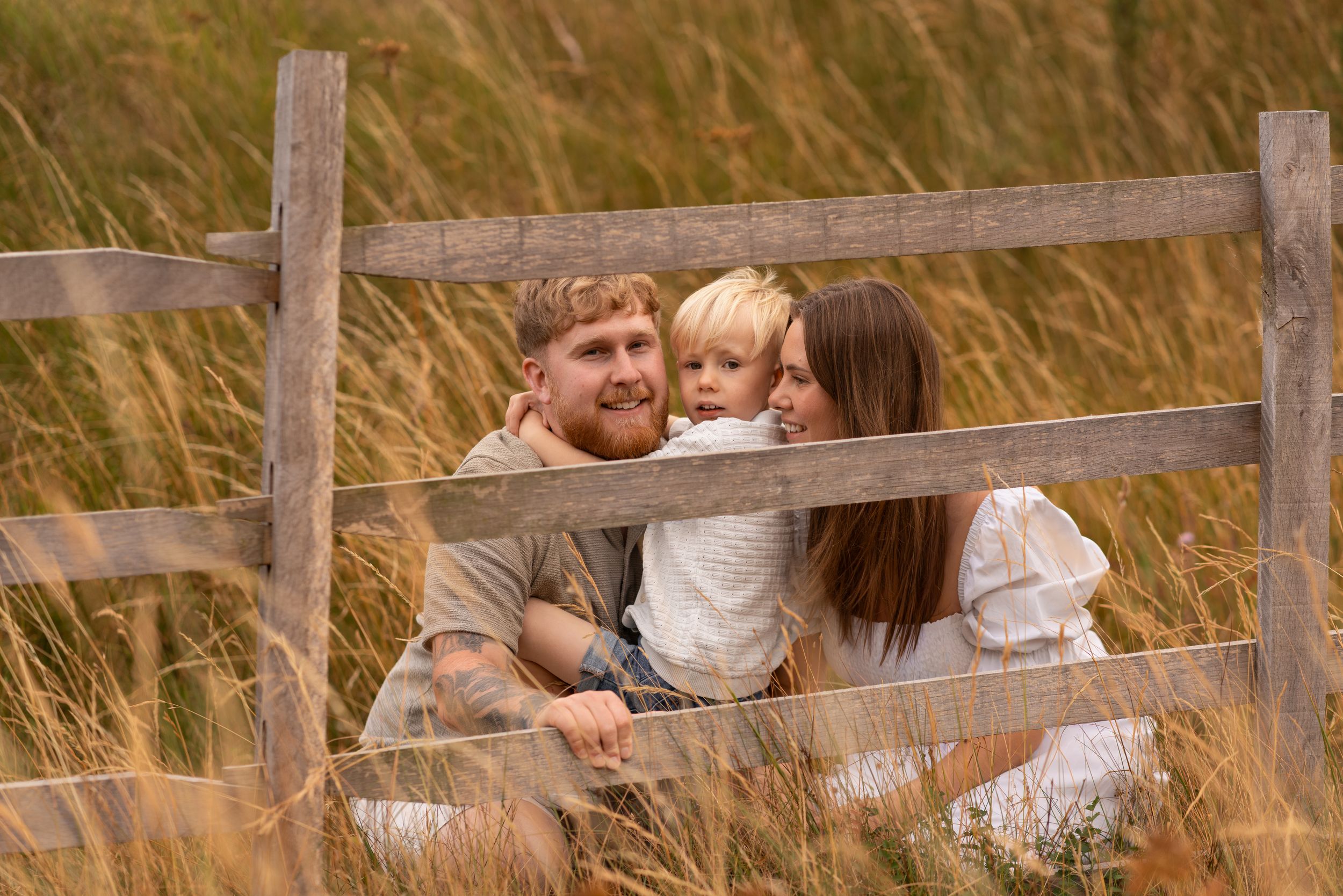 A happy family of three sitting together by a wooden fence in a golden grass field.