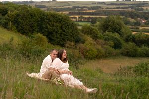 Couple sitting together in lush green countryside meadow, wearing cream outfits, enjoying scenic rural landscape view.