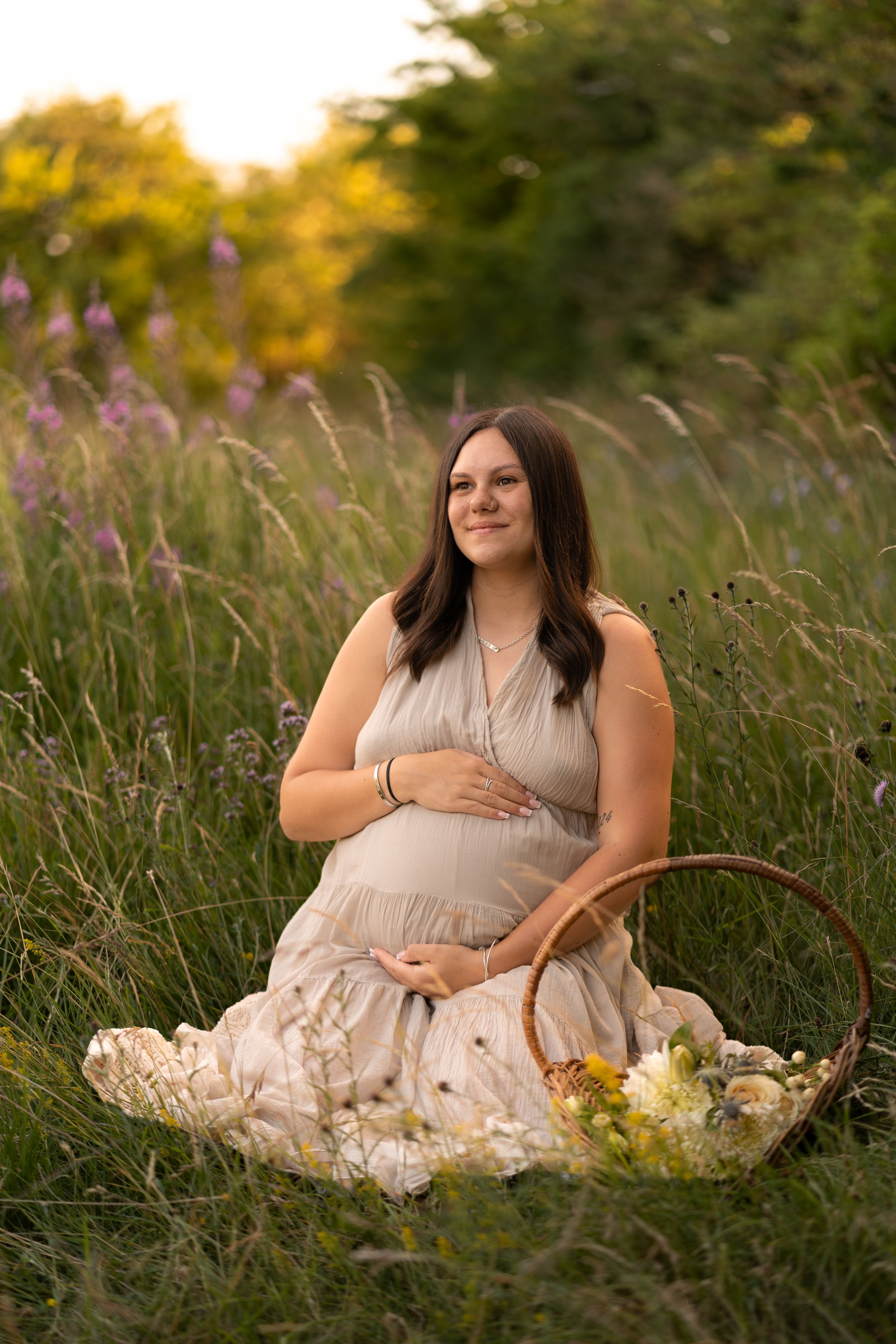 Pregnant woman in beige dress sitting in a green field with a flower basket during golden hour maternity photoshoot.