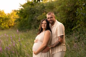 Pregnant couple poses in a sunlit meadow with wildflowers, woman in white dress cradling baby bump, man in beige shirt beside her.