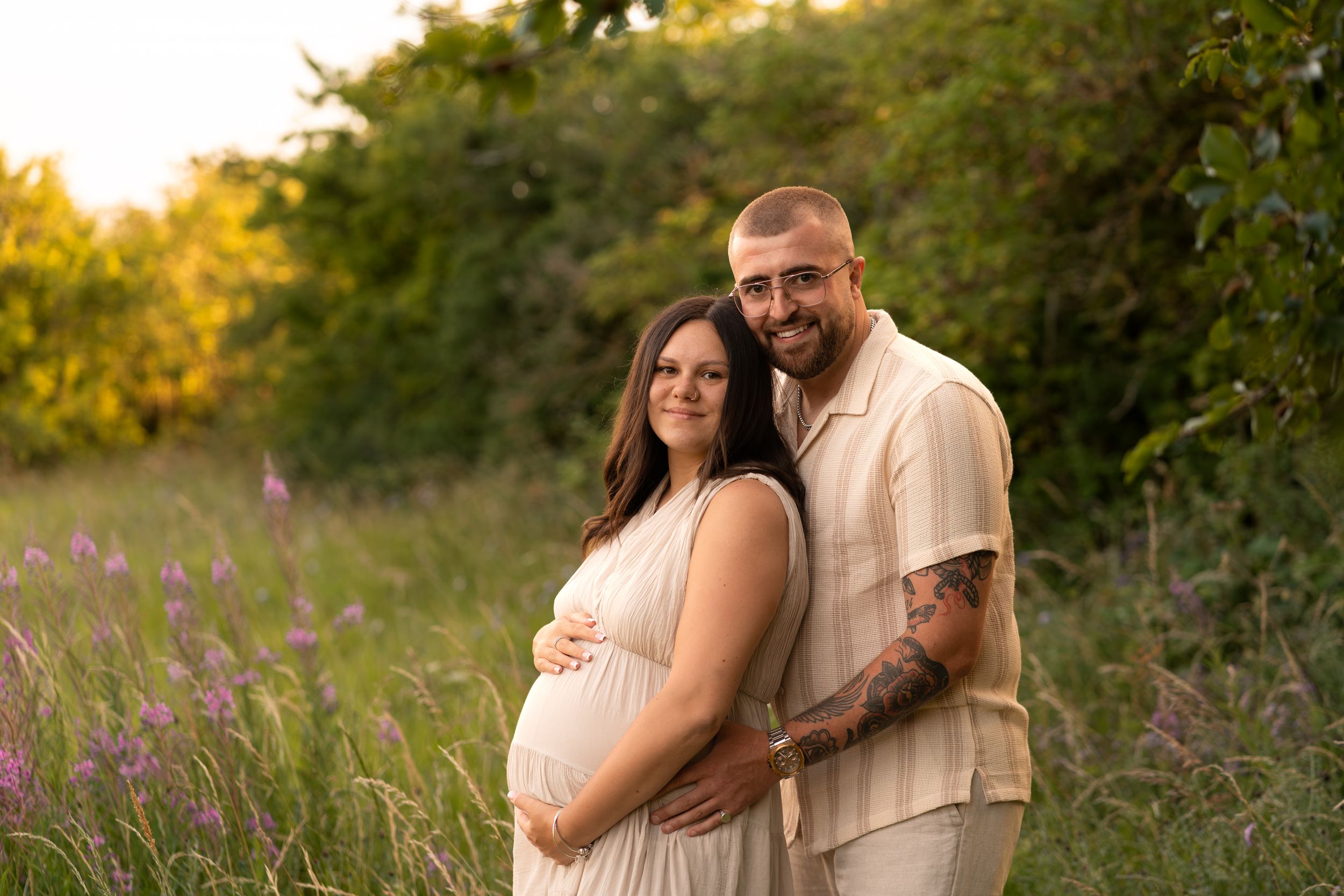 Pregnant couple poses in a sunlit meadow with wildflowers, woman in white dress cradling baby bump, man in beige shirt beside her.