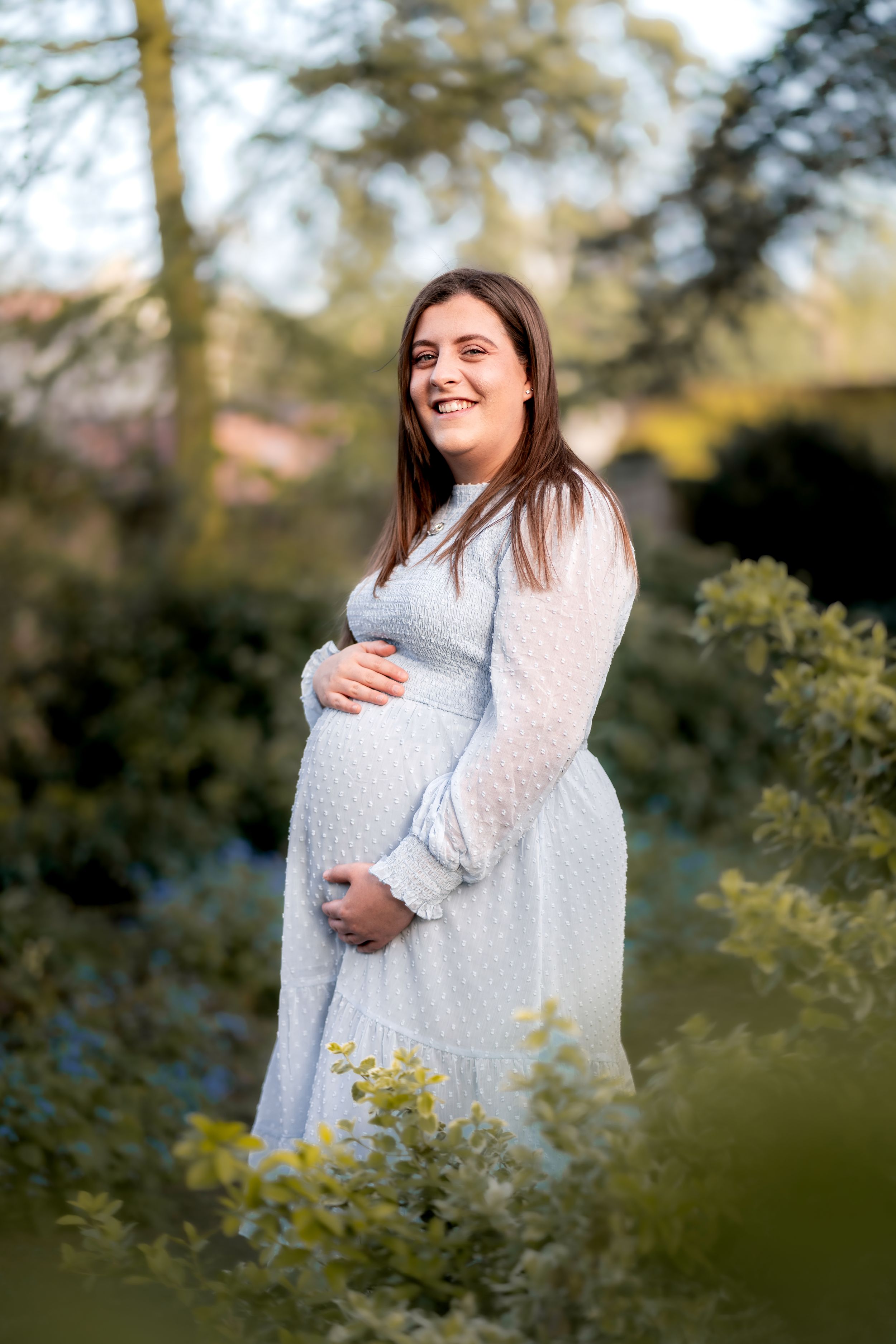 Smiling pregnant woman in a light blue dress cradling her baby bump while standing outdoors among green garden foliage.