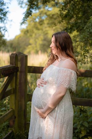 Pregnant woman in white lace off-shoulder gown cradling her baby bump outdoors by a wooden fence.