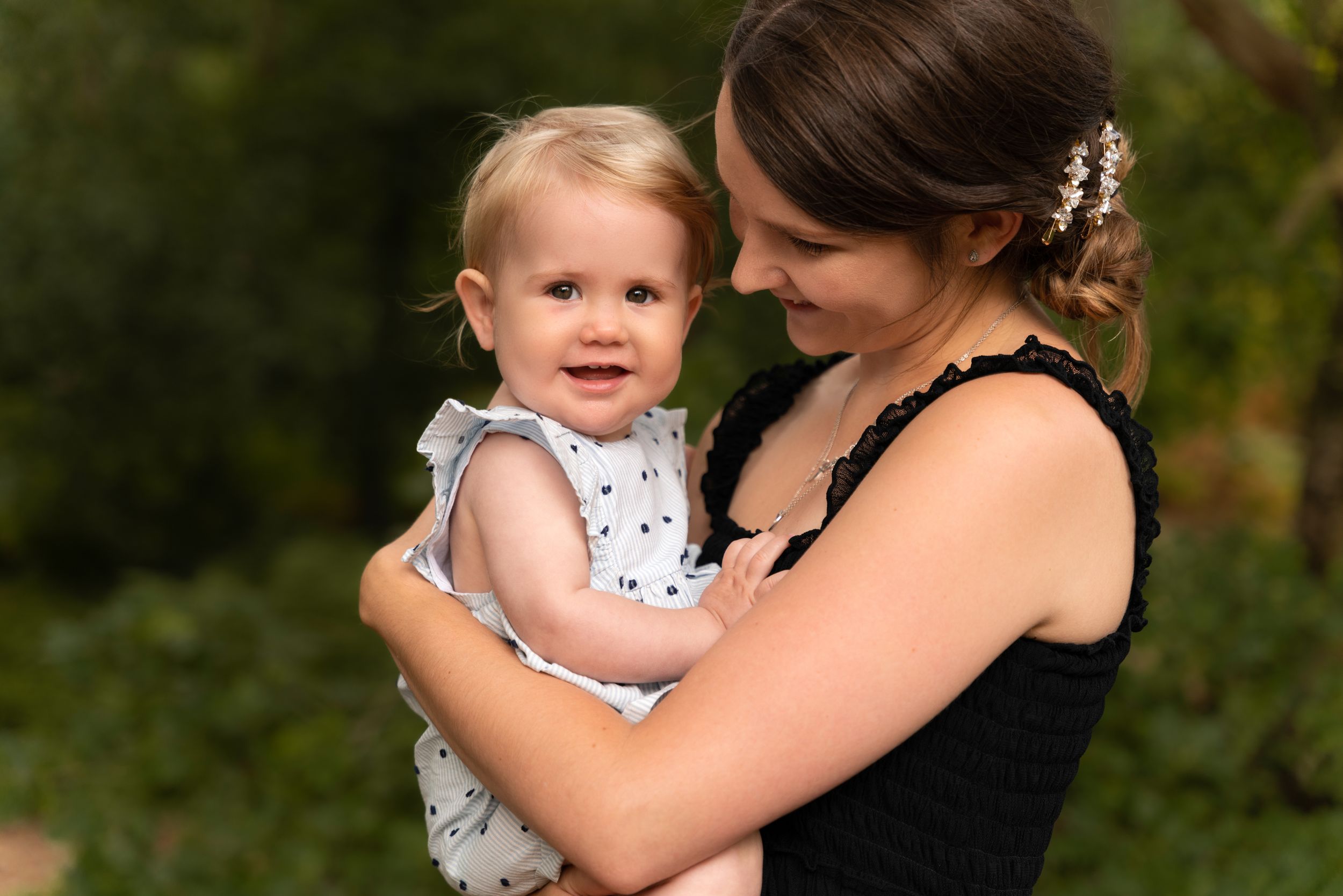A smiling mother in a black dress holds her happy blonde toddler girl outdoors during a family photoshoot in Lincolnshire.