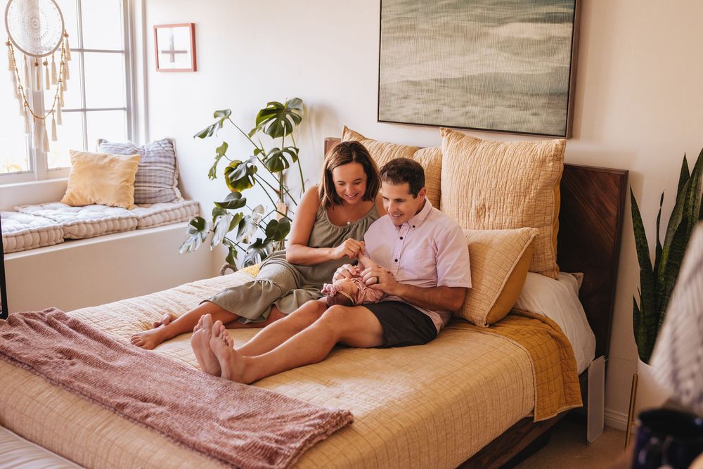 Couple sitting on bed with newborn baby in cozy bedroom with plants and natural light.