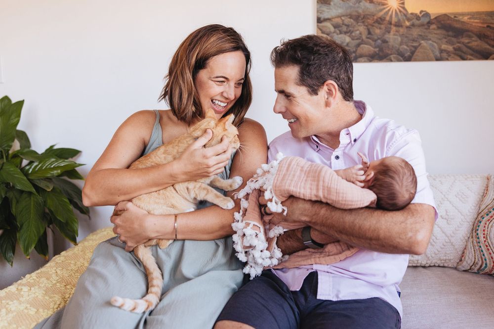 Happy couple sitting on couch holding newborn baby and orange tabby cat in bright modern home.