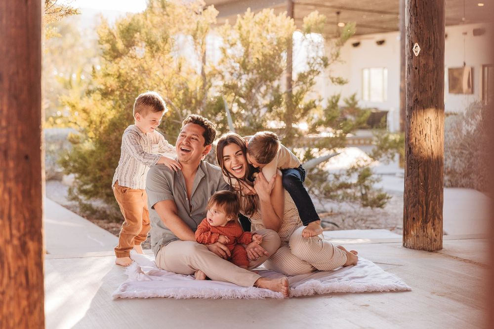Happy family with two parents and three young children sitting together on a blanket on a sunny outdoor patio.