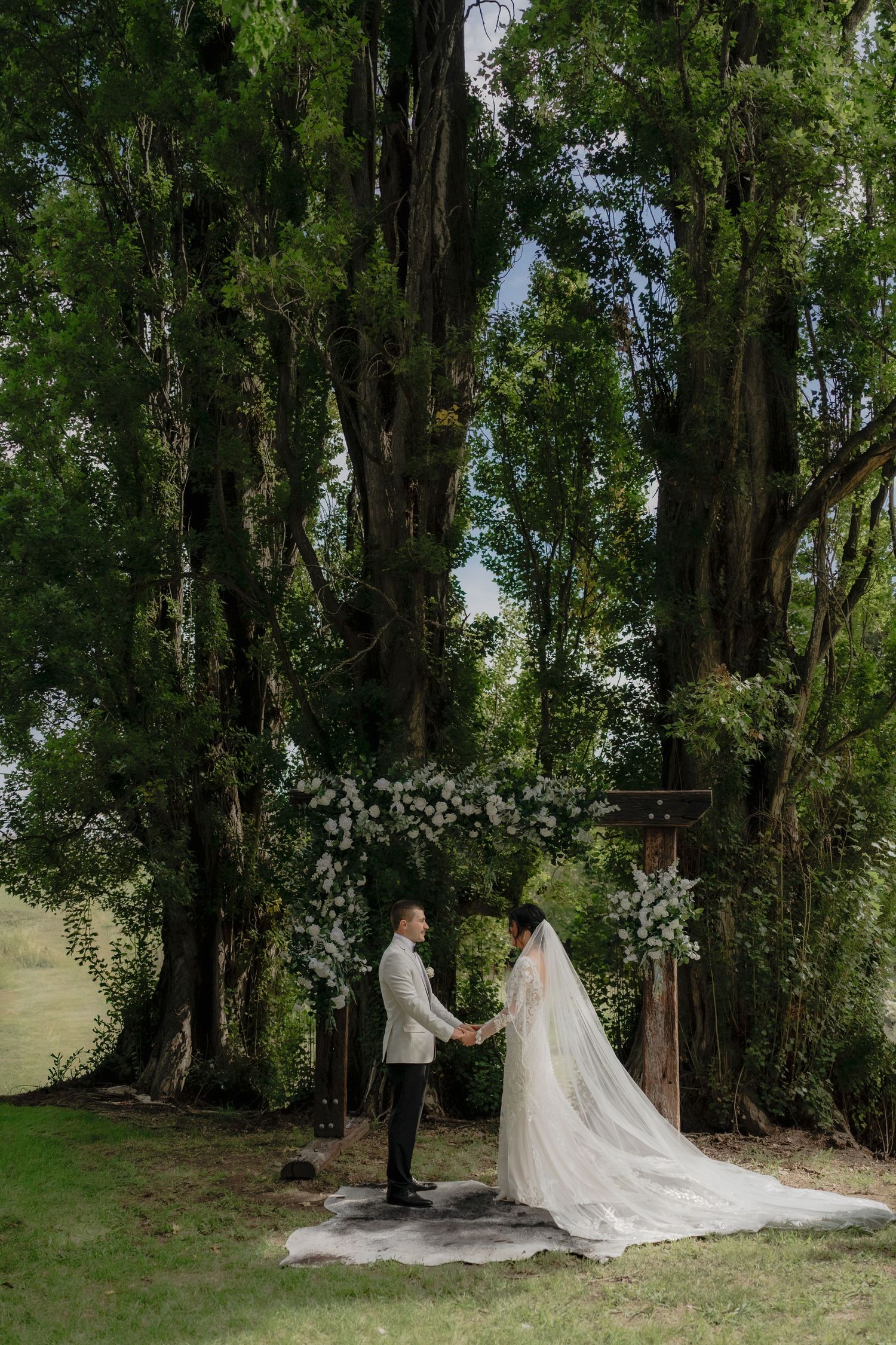 Bride and groom exchange vows under a floral arch surrounded by towering trees in a lush outdoor wedding ceremony.