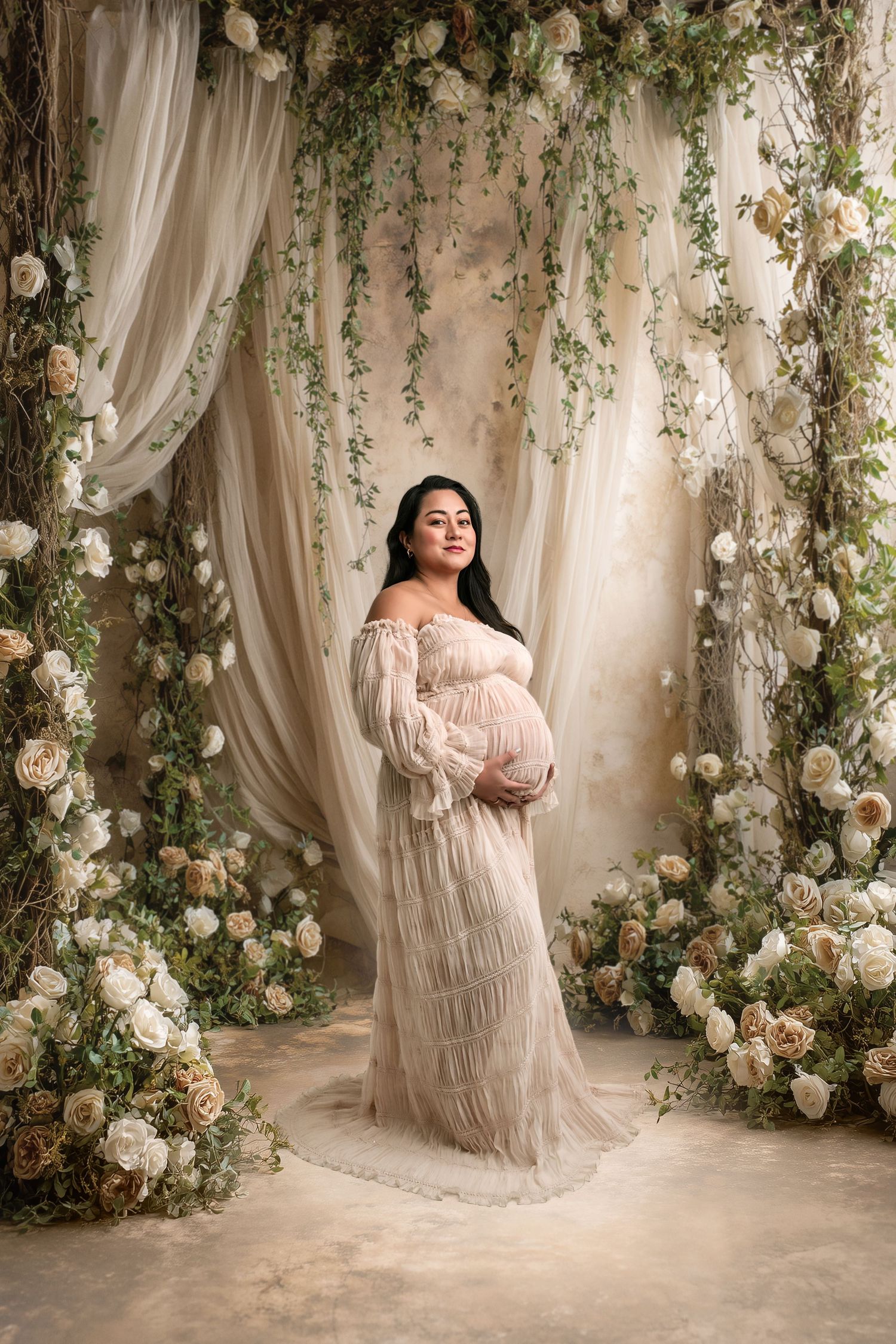 Pregnant woman in flowing beige lace gown poses among white roses and greenery in elegant maternity portrait on digital backdrop.