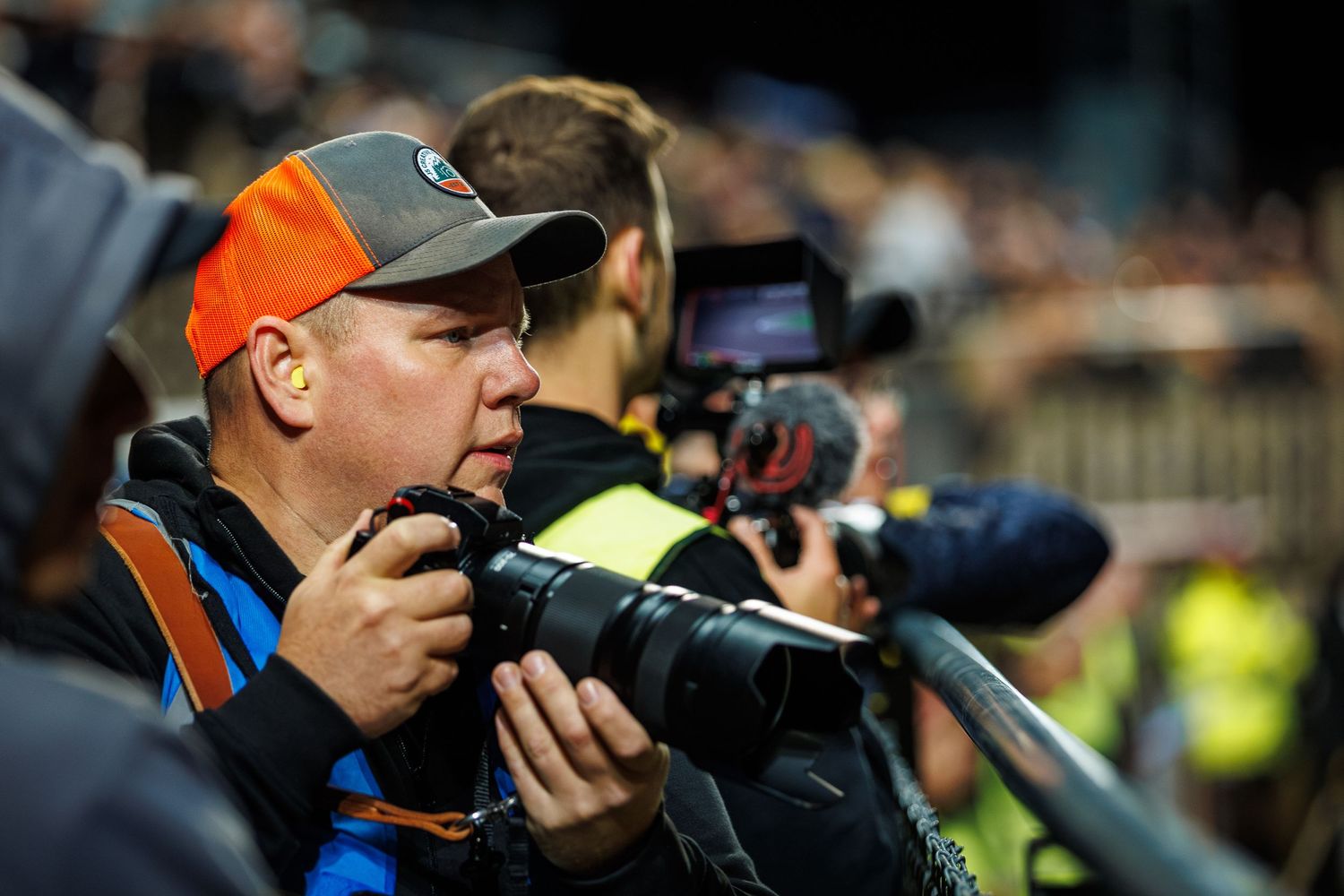 Fotograf Jonas Svensson står tillsammans med flera andra fotografer vid räcket runt en speedwaybana. Jonas är otroligt fokuserad på matchen.