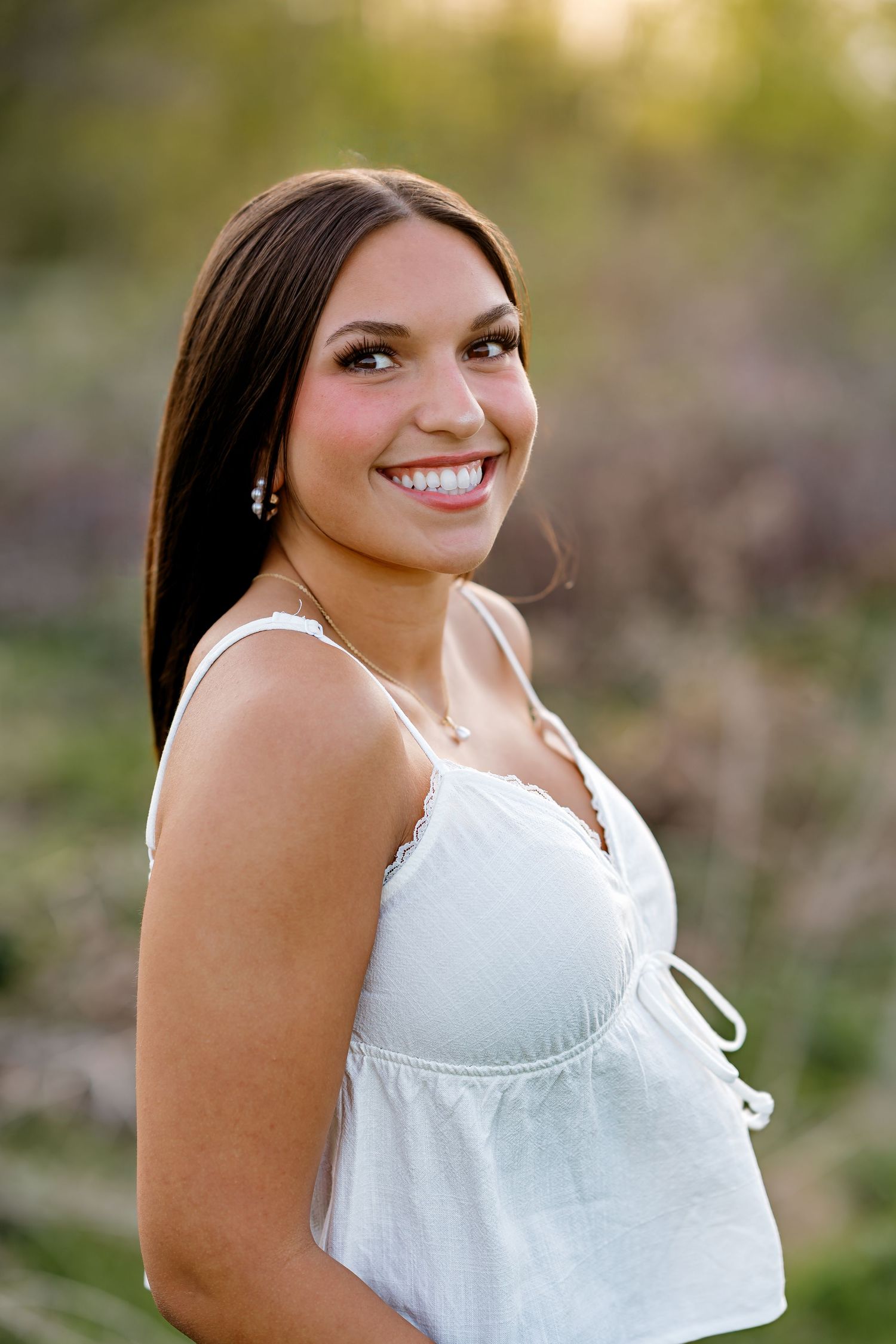 Young woman with long dark hair smiling outdoors in white sundress, golden hour bokeh background.