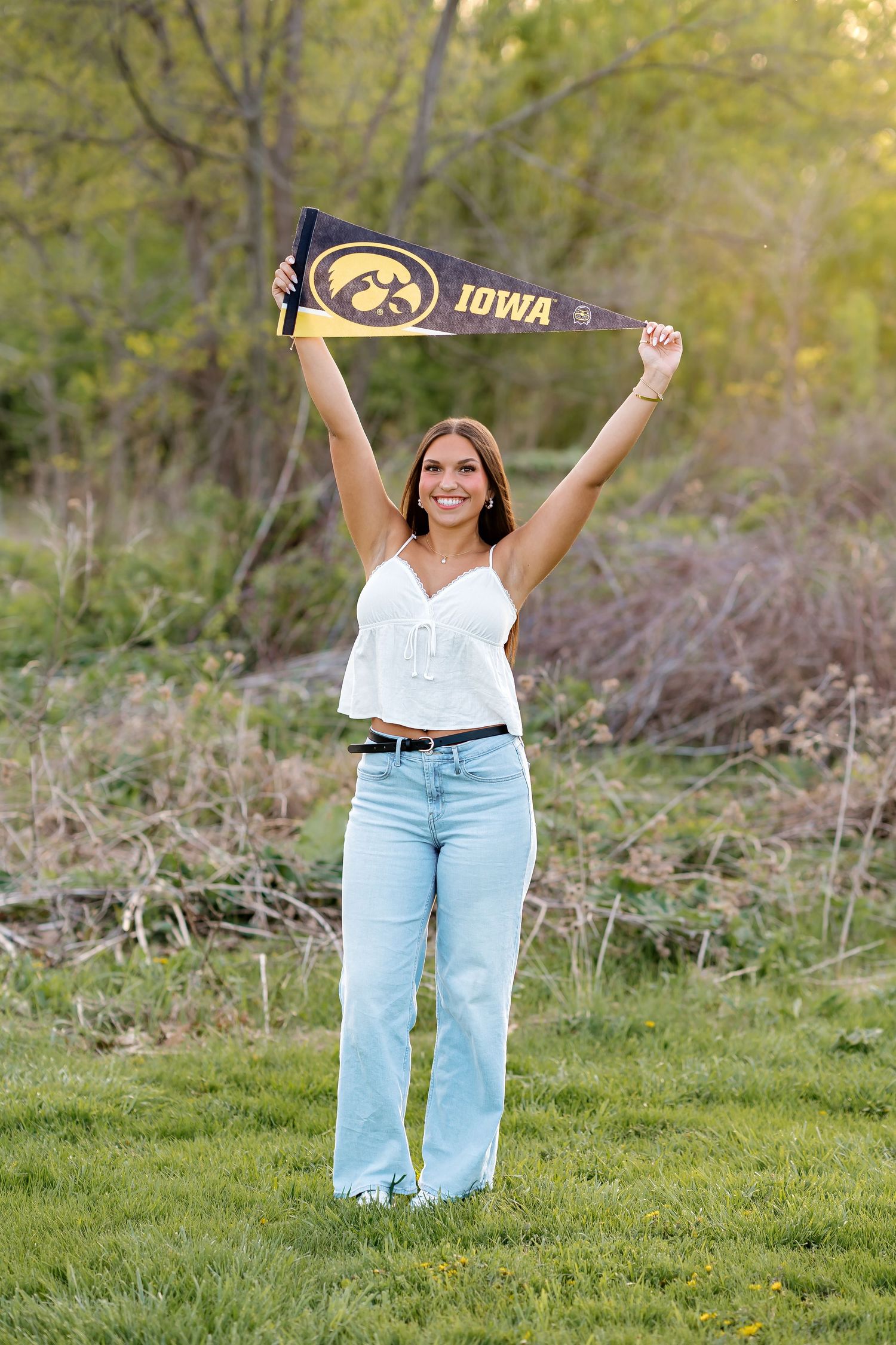 A smiling young woman holds an Iowa Hawkeyes pennant above her head outdoors in a green field.