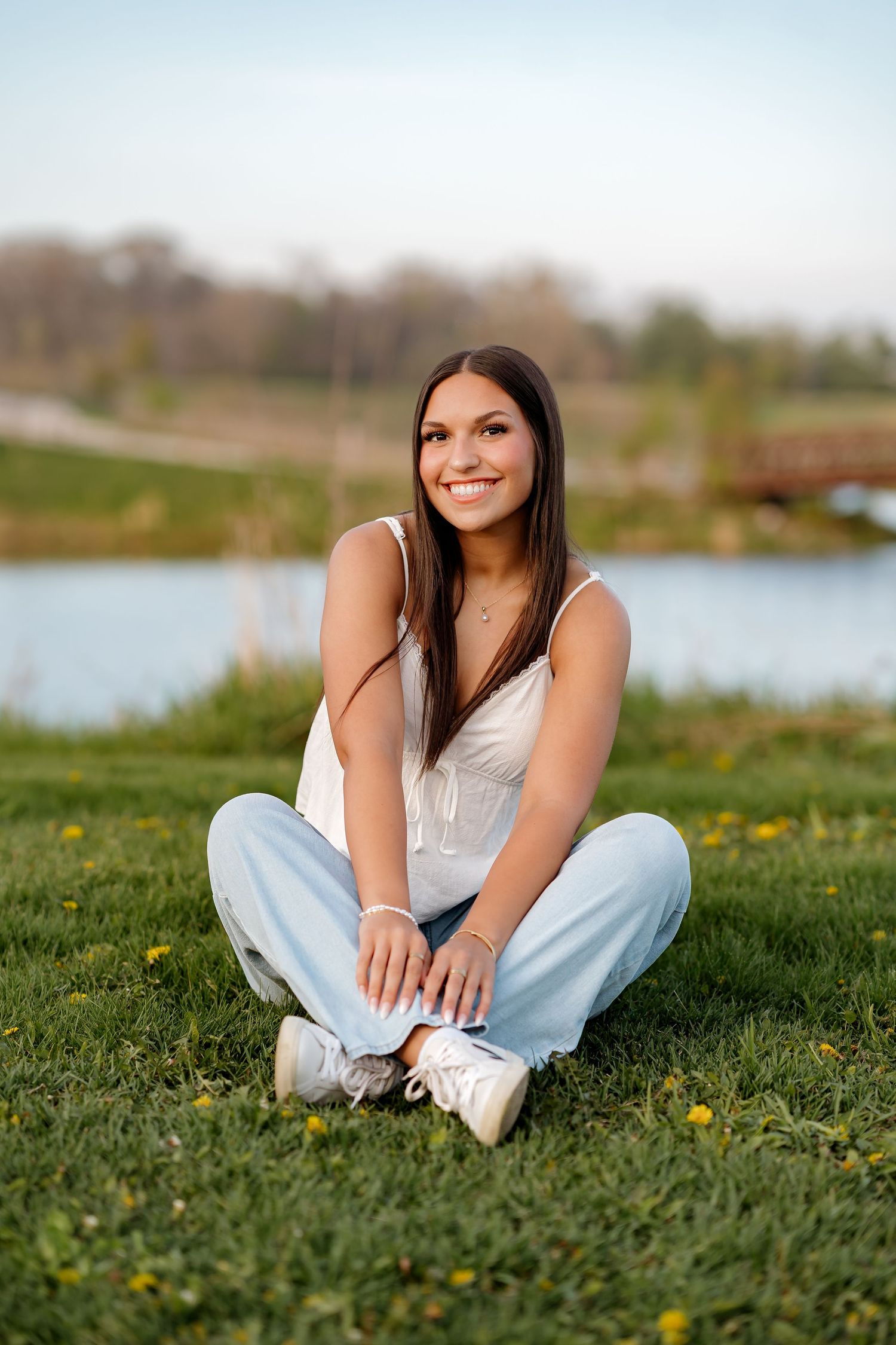 Young woman with long dark hair sits cross-legged on green grass, smiling, wearing white tank top and jeans near a pond.