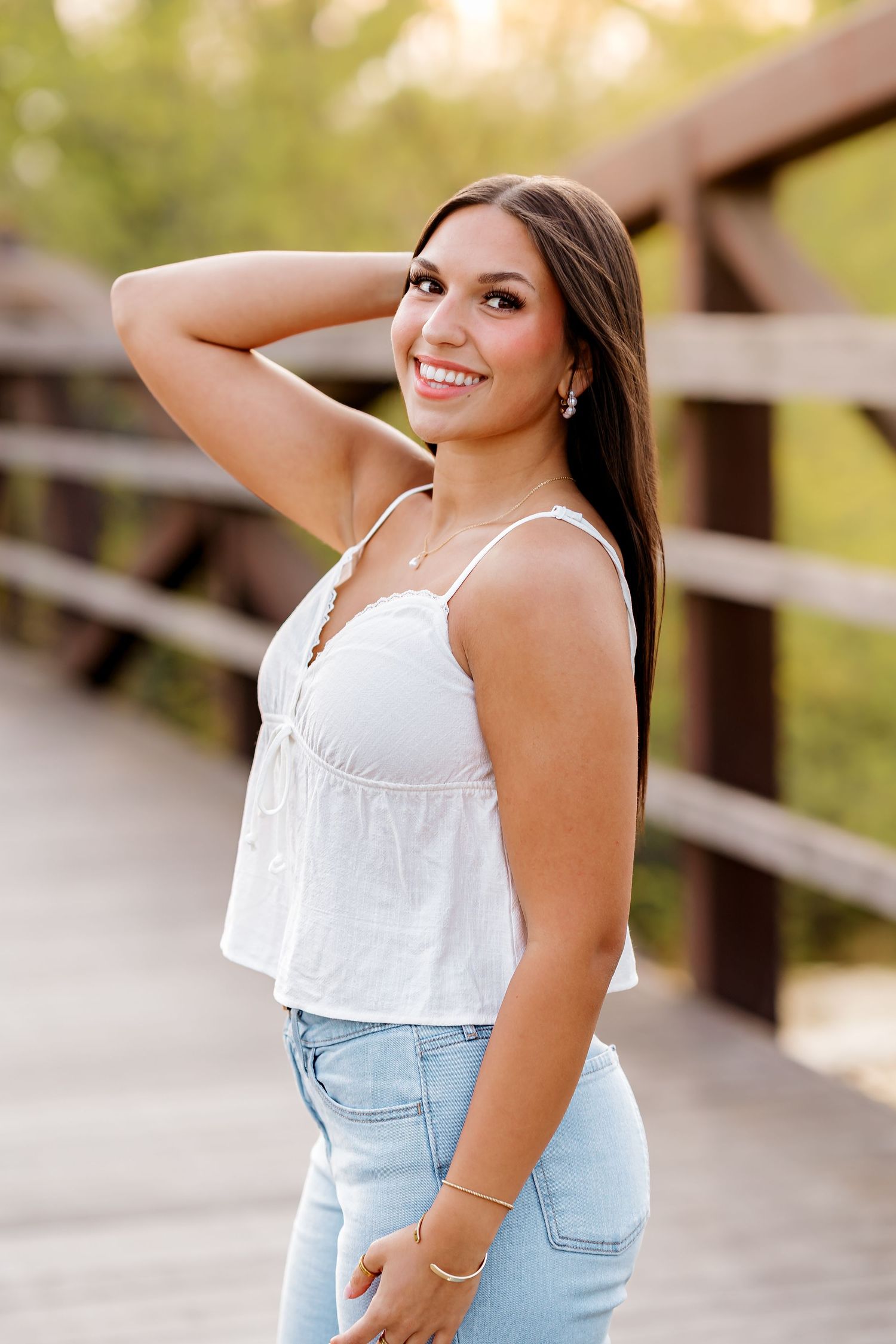 Young woman in white tank top and jeans smiling on a wooden bridge in golden hour light.