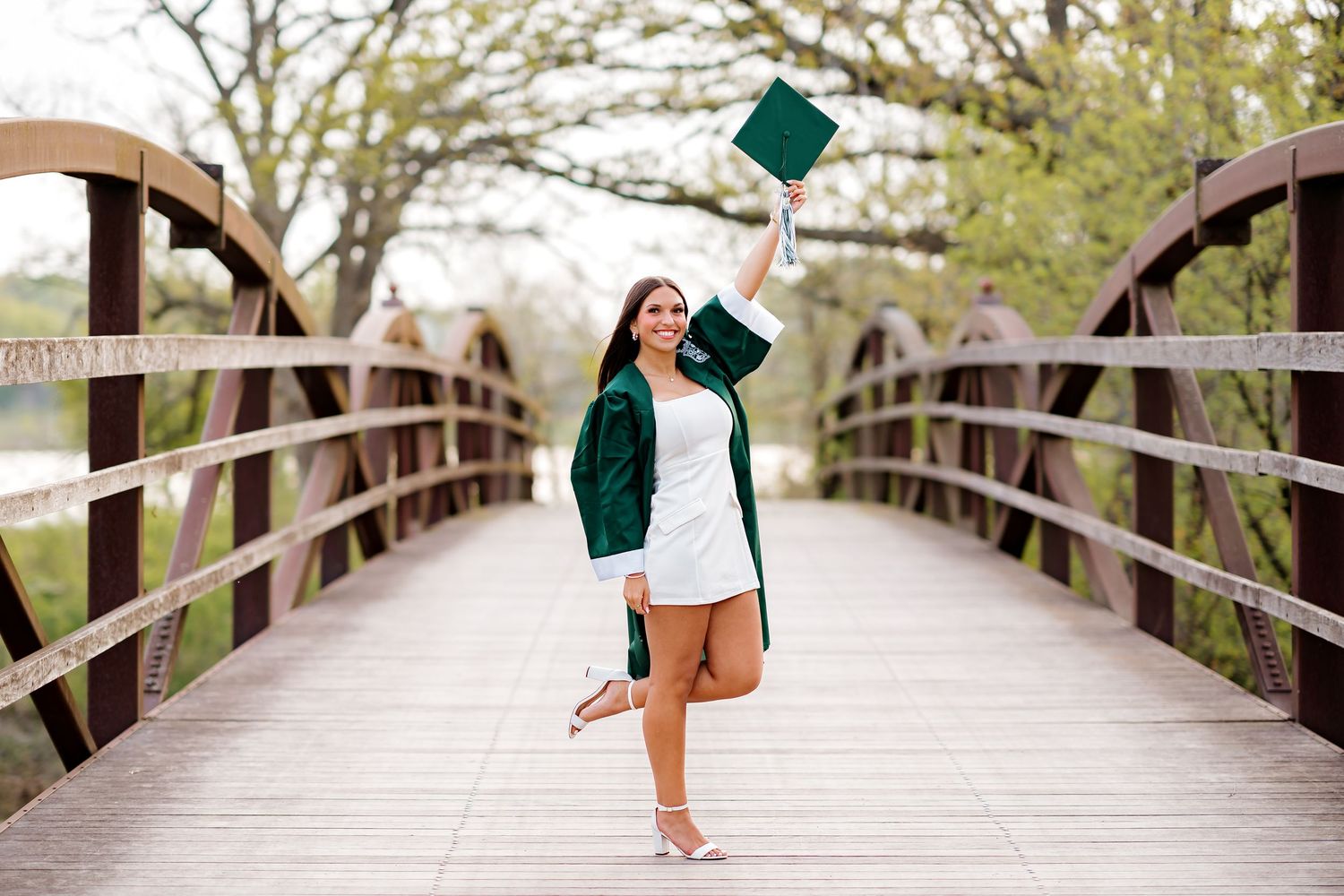 Graduate in green cap and gown celebrates on a wooden bridge, raising diploma while posing outdoors.