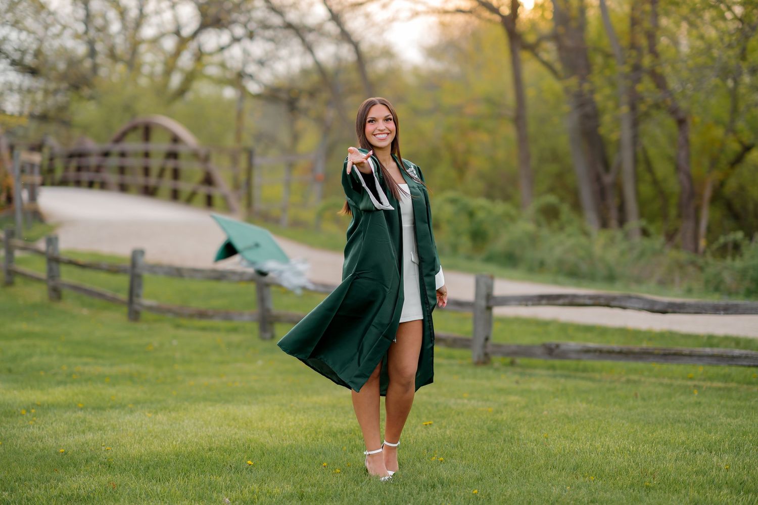 Young woman in green graduation gown smiles outdoors on lush lawn with wooden fence and bridge backdrop.