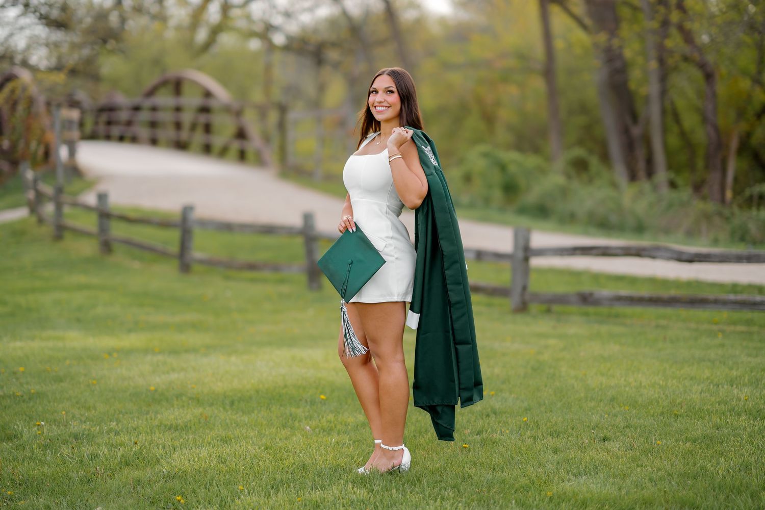 Young woman in white dress holding green graduation cap and gown, posing outdoors on green lawn near wooden fence.