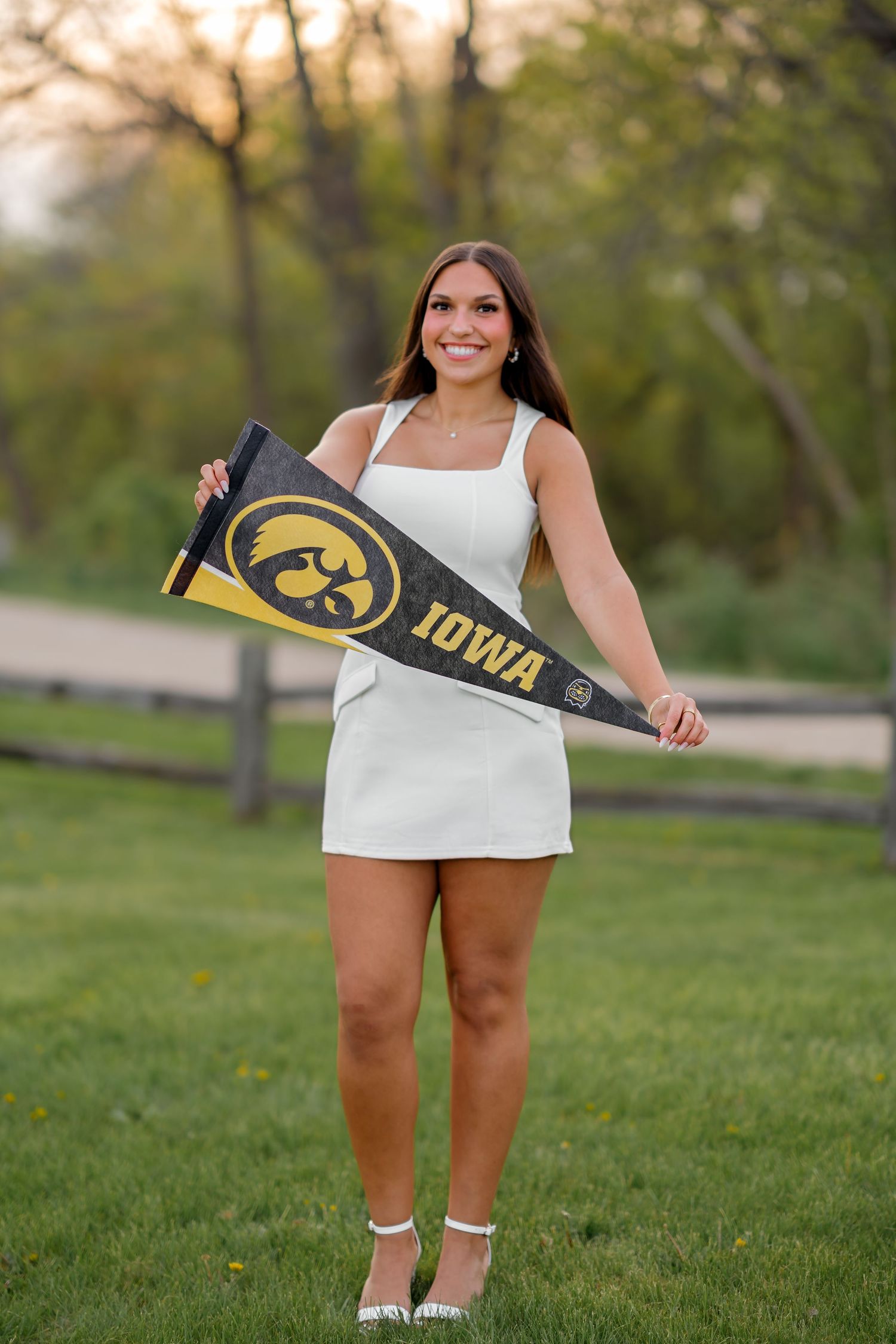 Smiling young woman in white dress holding an Iowa Hawkeyes pennant flag outdoors on green grass.