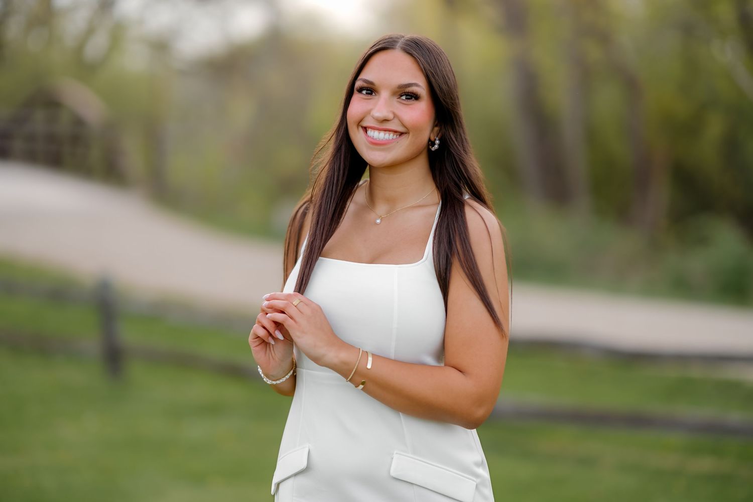 Young woman in white dress smiling outdoors on a scenic path with green trees and wooden fence.