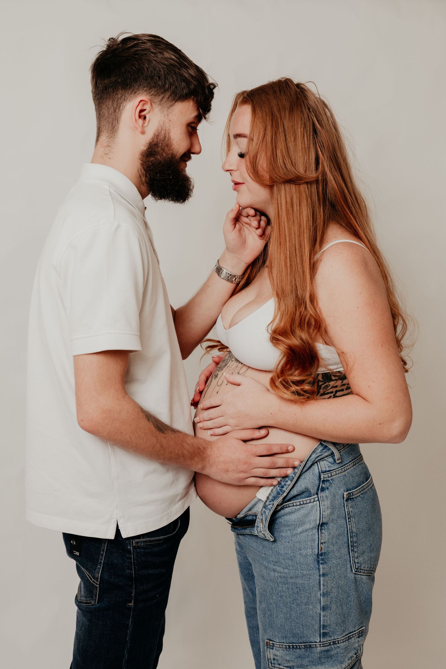 Bearded man in white shirt tenderly touches pregnant redhead woman's face while both hold her baby bump in studio.