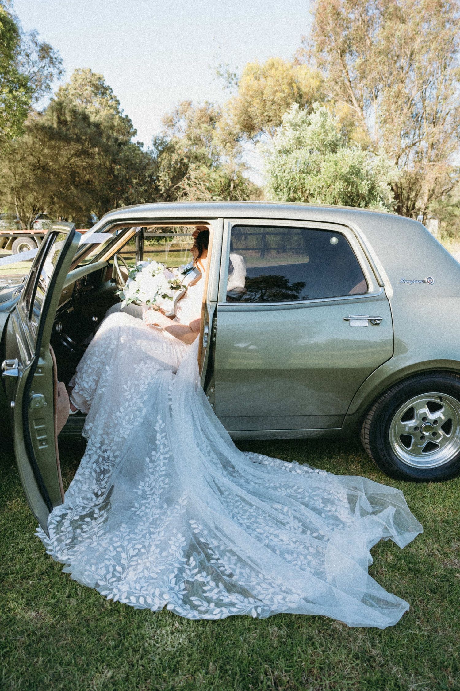 Bride in lace wedding gown with long train sits in vintage green car holding white bouquet outdoors.